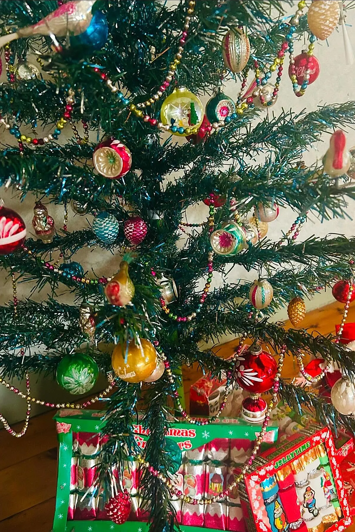 Vintage Christmas tree adorned with colorful glass ornaments in red, green, gold, and blue, including miniature scenes inside hollow ornaments and multicolored beaded garlands across dark green branches with wrapped holiday crackers underneath.