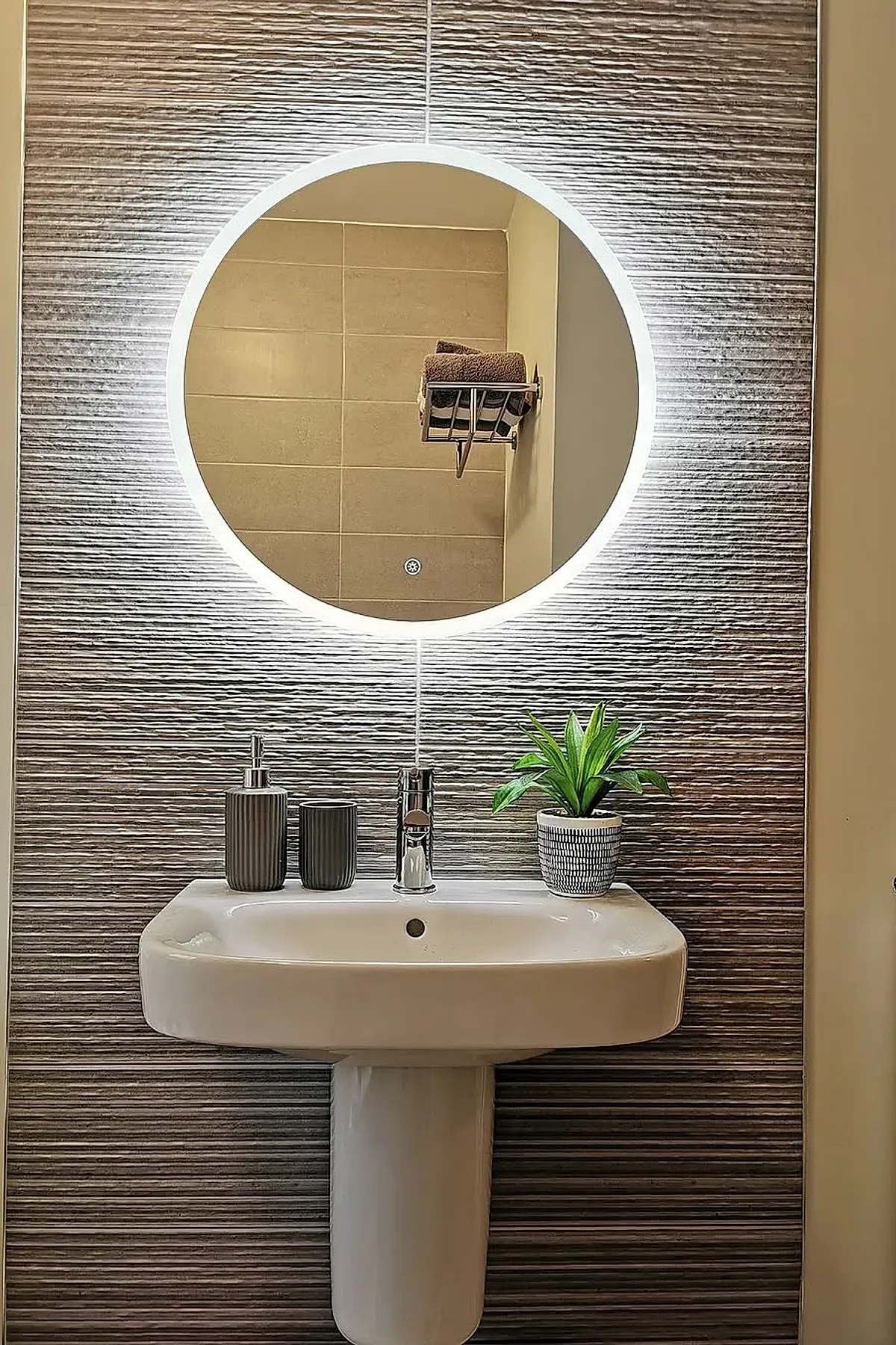 Small modern bathroom featuring a large round backlit mirror mounted on textured gray wall tiles, above a white pedestal sink with silver faucet, accessorized with gray soap dispenser, tumbler, and small potted green plant.