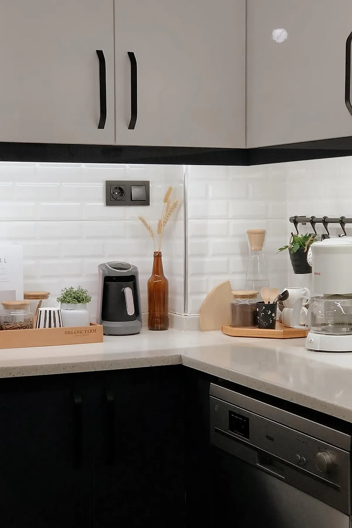 Minimalist kitchen countertop coffee bar ideas featuring white subway tile backsplash, beige and black coffee machines, wooden trays holding jars and cups, a small potted plant, dried grasses in a brown bottle, and hanging herbs on black hooks against light gray upper cabinets and black lower cabinets.