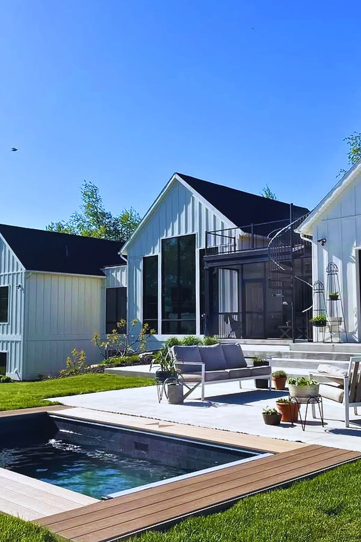 A small plunge pool set into a wooden deck in a modern backyard featuring white siding houses, concrete patio with gray cushioned outdoor furniture, potted plants, and bright green grass under clear blue sky.