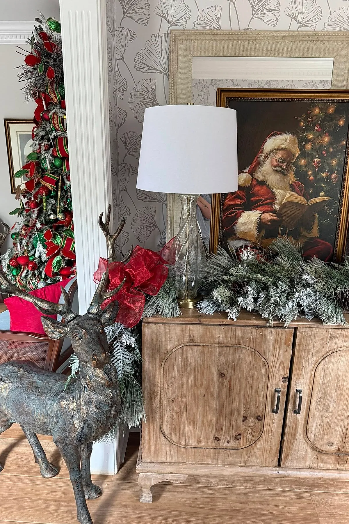 A wooden cabinet styled for Ralph Lauren Christmas with a frosted pine garland topped by a red bow, a clear glass table lamp with a white shade, a framed painting of Santa Claus reading, and a bronze deer sculpture beside a decorated Christmas tree in the background.