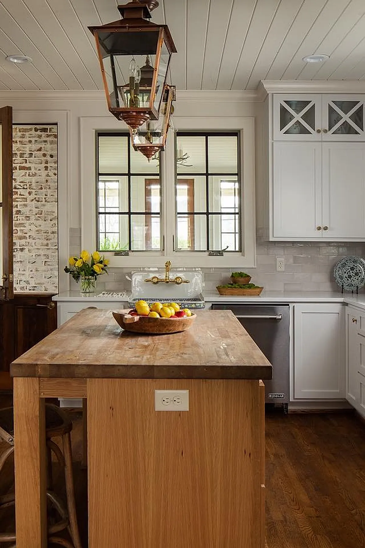 Cottage kitchen featuring white glossy subway tile backsplash behind a vintage-style white sink with an ornate gold faucet, wooden countertop island with a bowl of lemons and apples, white cabinetry with glass-front upper cabinets, large black-framed windows above the sink, and copper pendant lights hanging from a white paneled ceiling.