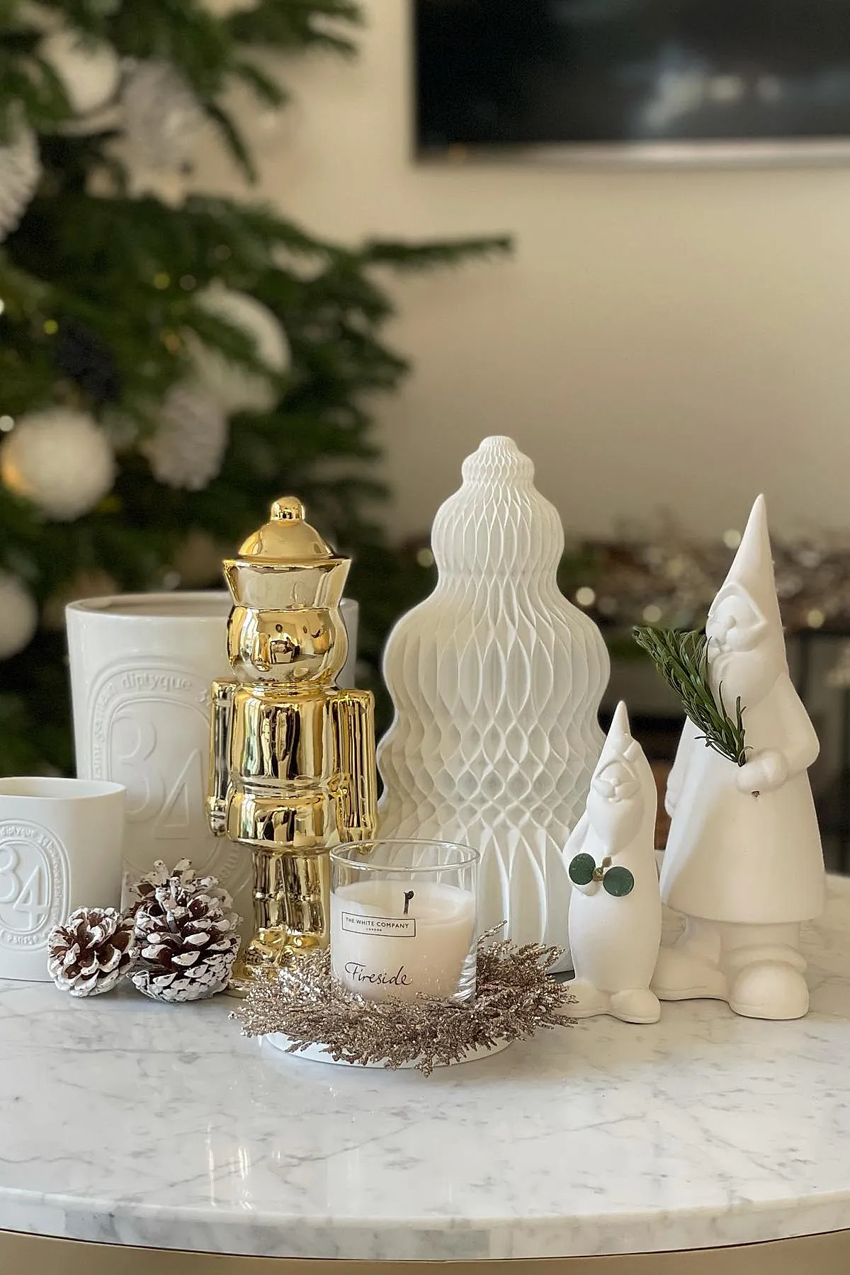 christmas coffee table decor featuring a reflective gold nutcracker figure, two white ceramic gnome statues holding greenery, a textured white paper tree, a lit candle labeled