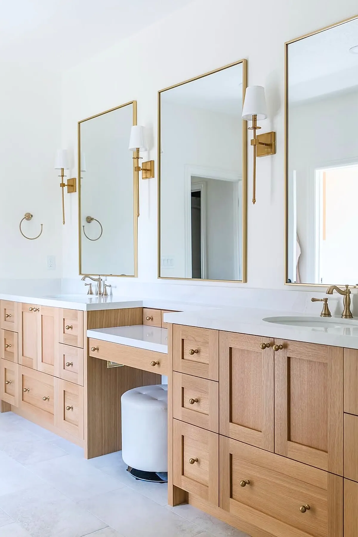 Master bathroom featuring light wood cabinetry with brass knobs, white countertops with dual sinks, three large gold-framed mirrors above each sink, brass wall sconces with white shades, and a white cushioned stool tucked under a vanity area.