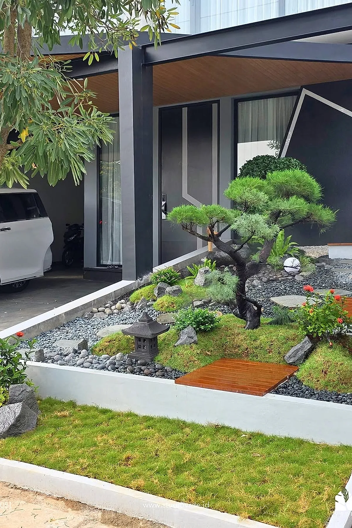 Small front yard landscaping with Japanese style garden featuring a bonsai tree, green moss-covered mounds, smooth gray pebbles, stone lantern, wooden stepping path, and neatly trimmed shrubs in front of a modern house with black and gray exterior.