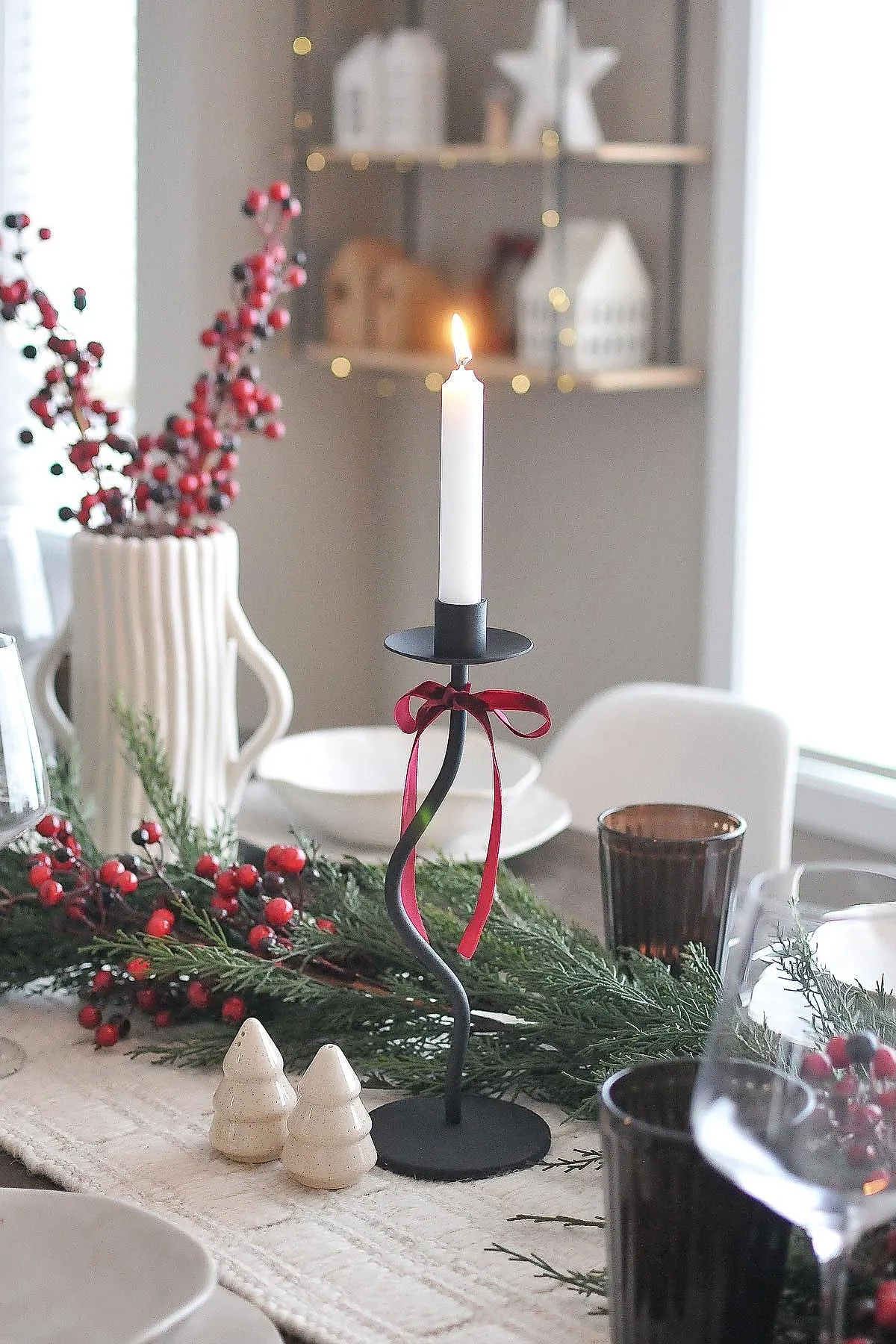 minimalist christmas dinner table settings featuring a black twisted metal candleholder with a white taper candle tied with a red ribbon, greenery garland with red berries, cream ceramic tree salt and pepper shakers, dark glassware, and white dishes on a textured beige table runner.