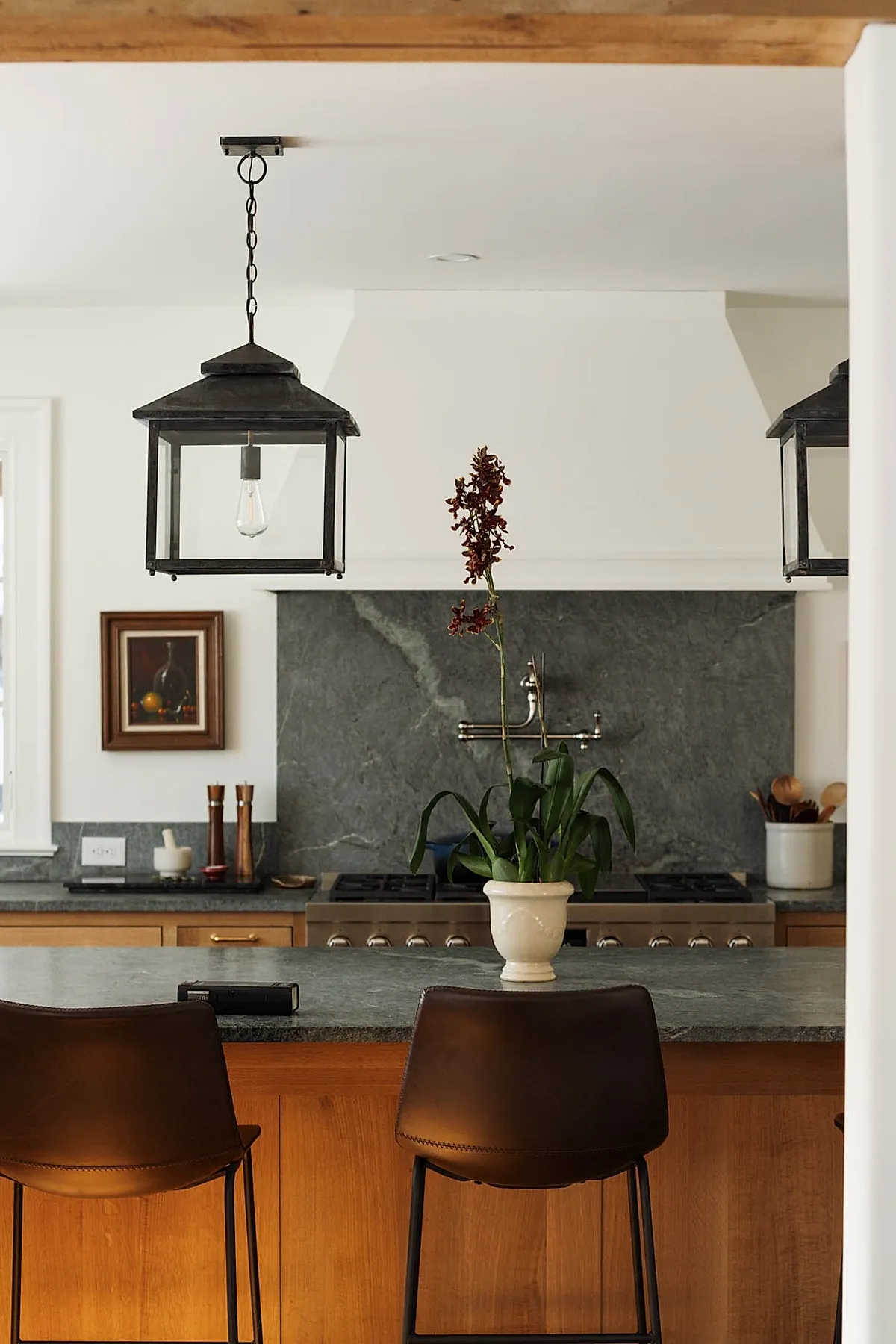 Rustic kitchen featuring a dark gray natural stone backsplash behind a stove, wooden cabinetry with brass handles, two black lantern-style pendant lights hanging from the ceiling, and a potted plant in a cream ceramic vase on the countertop.