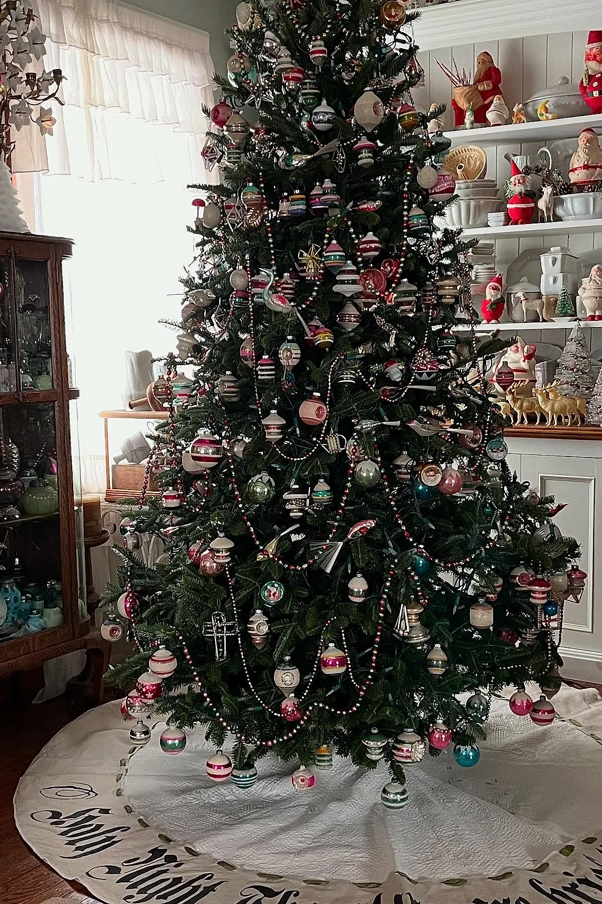 Vintage Christmas tree decorated with striped glass ornaments in red, white, green, pink, and blue hues, draped with red and black beaded garlands, standing on a white tree skirt embroidered with