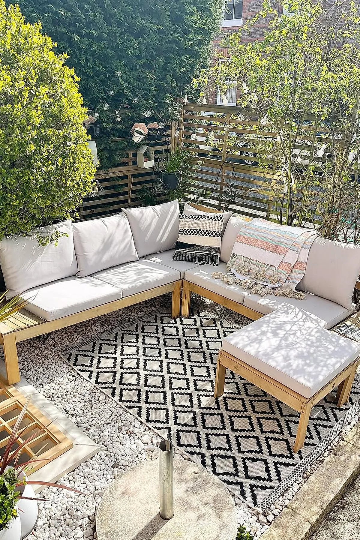 Small front yard landscaping featuring a cozy L-shaped wooden outdoor sofa with beige cushions, patterned black and white rug, light-colored pebbles surrounding the seating area, wooden slatted fence with string lights, and lush green plants providing privacy.