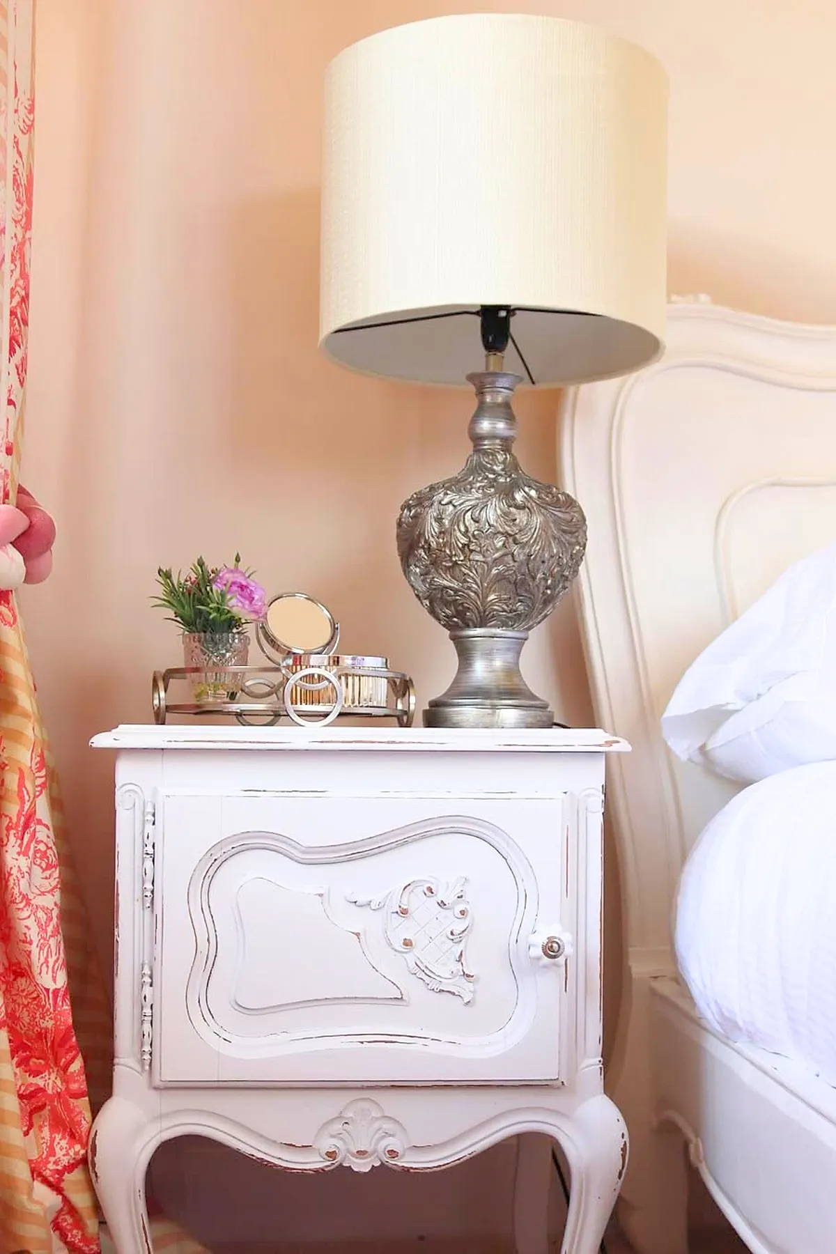 A white distressed wooden nightstand with ornate carvings holds a large vintage silver lamp with intricate leaf patterns and a cream-colored lampshade, next to a bed with a white upholstered headboard and soft peach walls in a shabby chic bedroom.