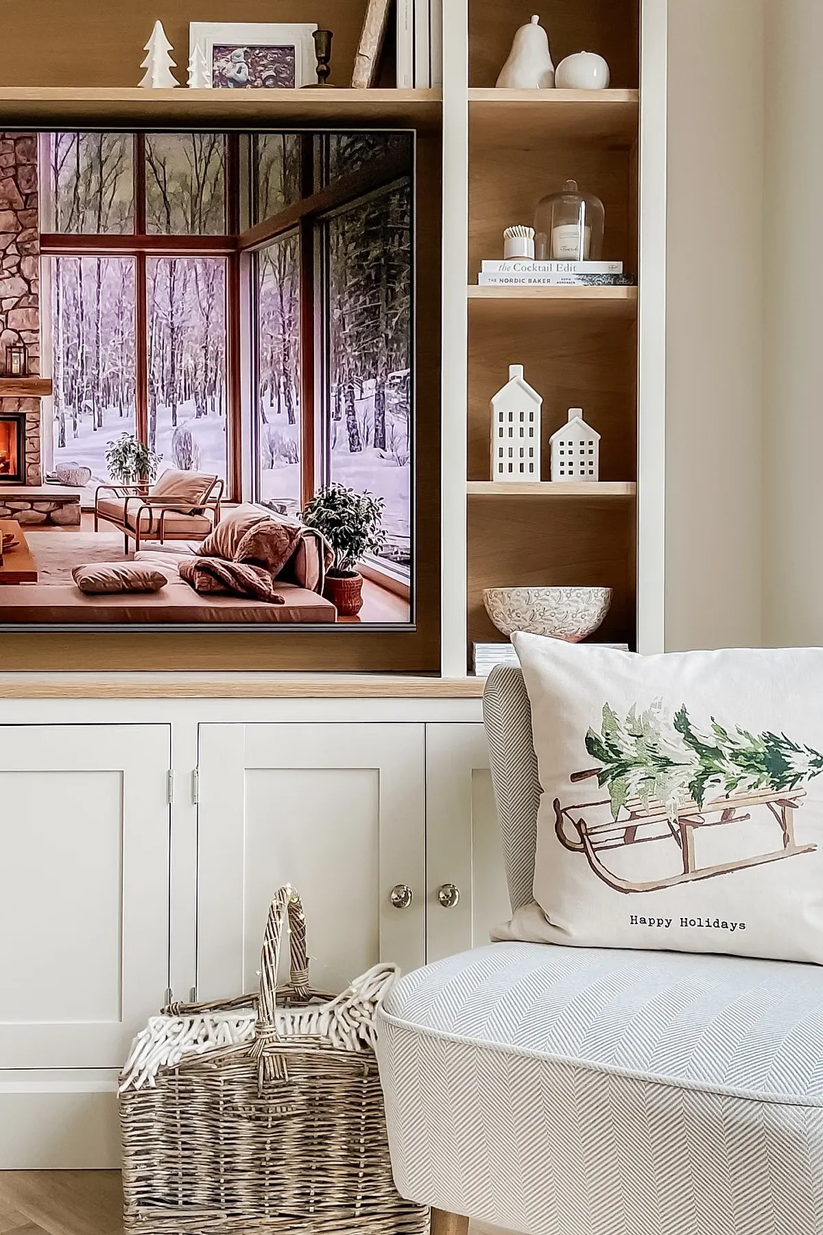 neutral minimalist christmas decor featuring built-in wooden shelves with white ceramic houses and pears, a wicker basket with a cozy throw, and a light grey chair adorned with a white pillow printed with a sleigh carrying pine trees
