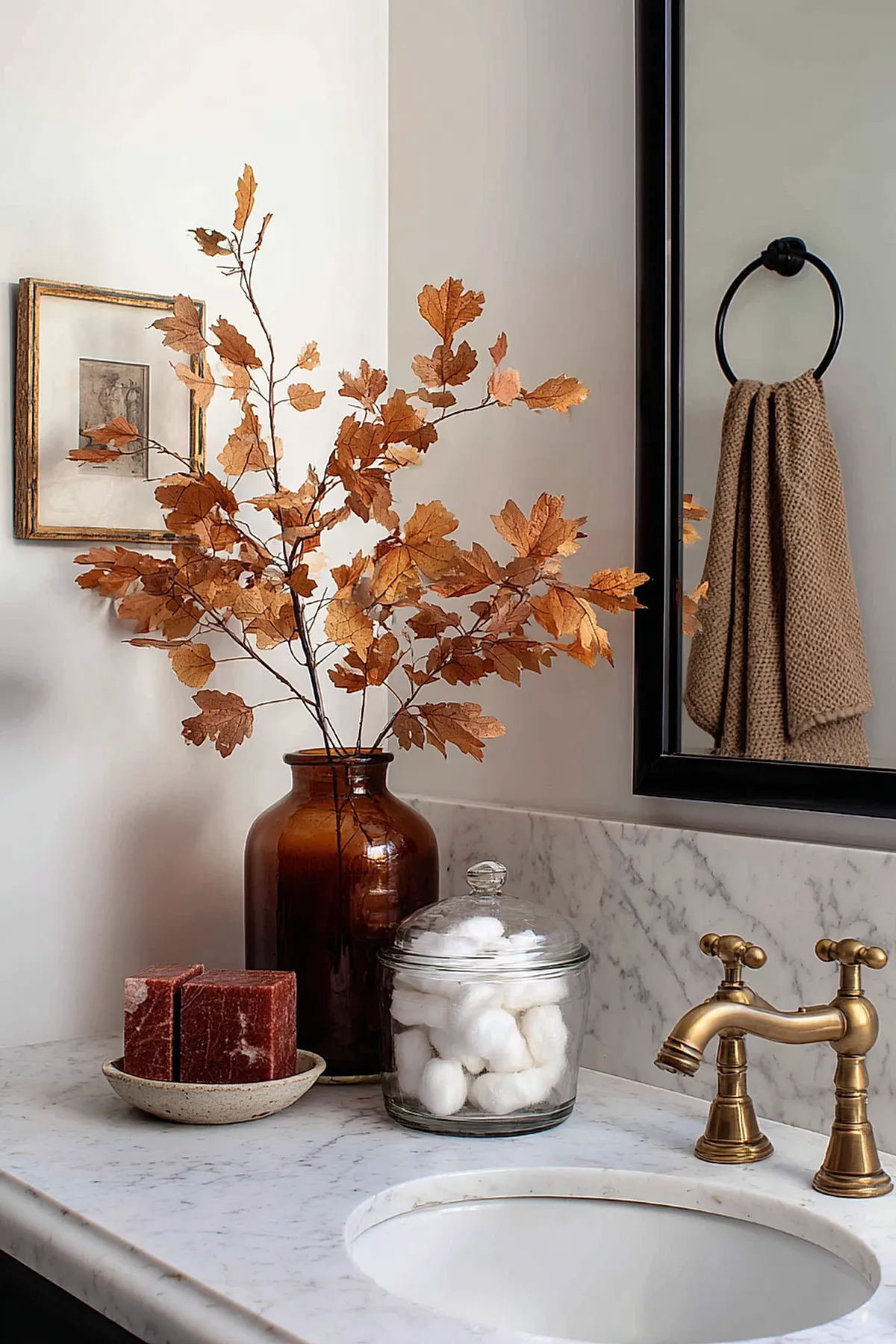 marble bathroom countertop with amber glass vase holding branches of dried brown fall leaves, red soap bars in ceramic dish, glass jar with cotton balls, brass faucet, black towel ring holding brown knit towel