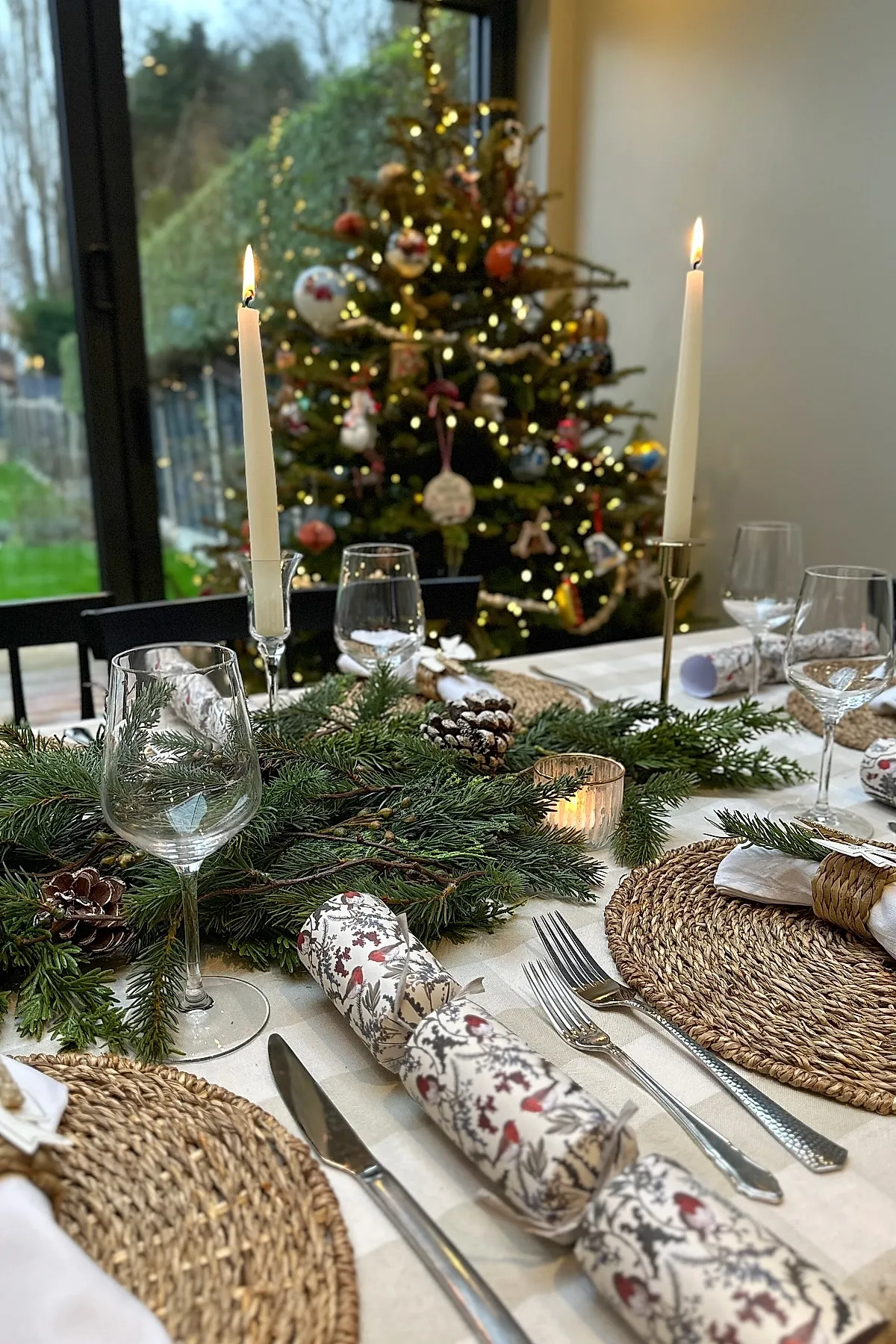 christmas dinner table settings featuring natural evergreen branches, pinecones, woven round placemats, white napkins with wicker rings, patterned Christmas crackers, clear wine glasses, silver cutlery, and tall white taper candles with a decorated Christmas tree in the background.