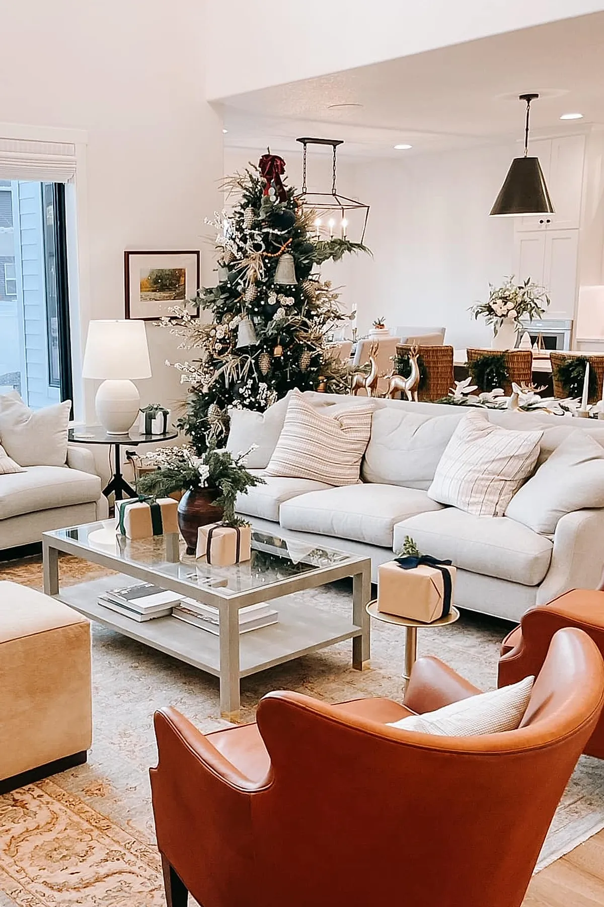 christmas living room decorated with a green tree adorned with white ornaments and natural accents, beige sofas with striped pillows, brown leather armchairs, glass coffee table holding wrapped gifts and greenery in a brown vase, cream walls, and woven dining chairs with gold reindeer figurines in the background