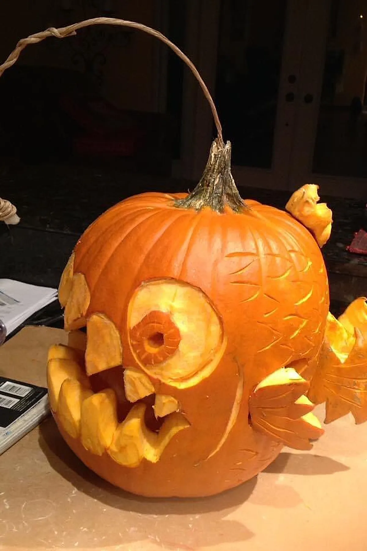 Orange pumpkin carved as an anglerfish with large round eyes, sharp triangular teeth, detailed etched scales, and a long curved stem acting as the fishing lure antenna.