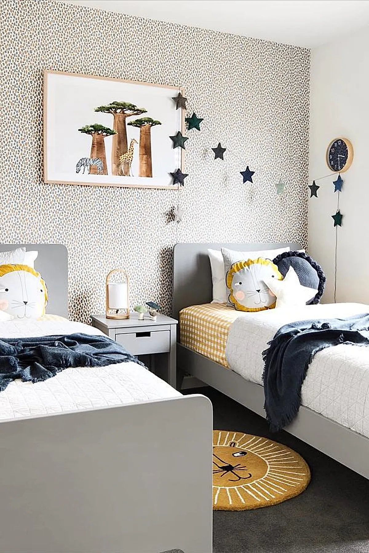 Two gray twin beds with white and yellow gingham bedding, lion face pillows, and dark blue throws. Beige wall with small irregular brown spots, framed artwork of trees and animals above beds, navy star garland draped along the wall near a round wooden clock.