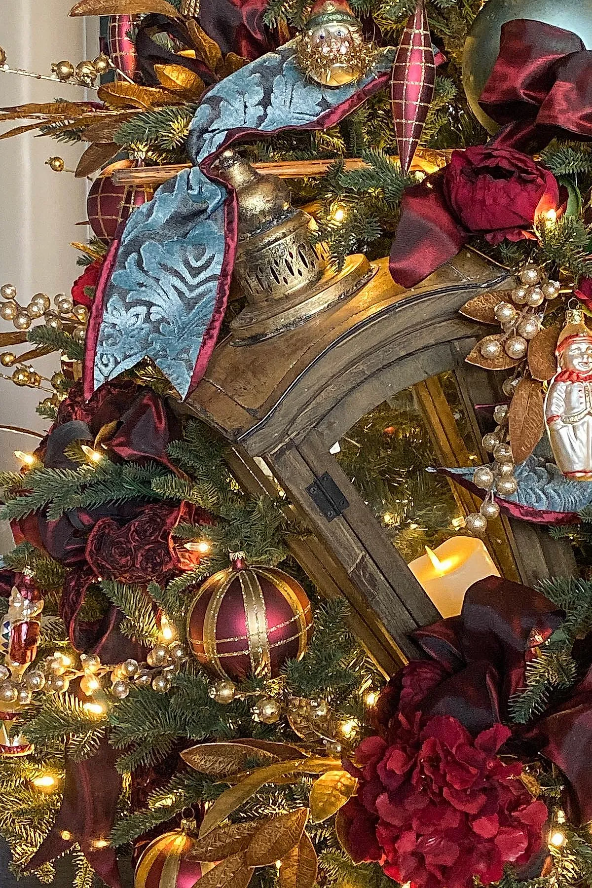 Close-up of a traditional christmas tree decorated with deep burgundy velvet ribbons, gold leaves, pearl berry clusters, red and gold striped baubles, vintage figurine ornaments, and an antique-style lantern holding a glowing candle nestled among green pine branches.
