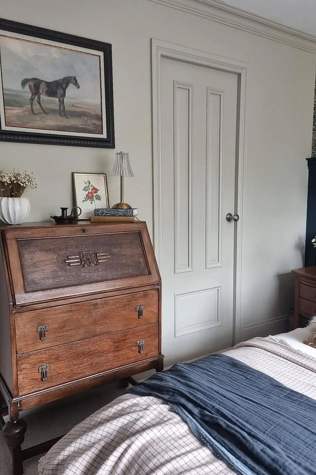 vintage bedroom corner featuring dark wood antique writing desk with metal hardware, framed horse painting on beige wall, small table lamp, books, dried flowers in white vase