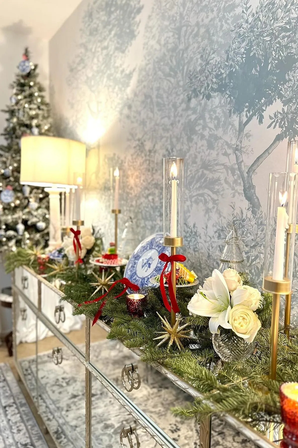 Elegant interior with a mirrored console table, candles, blue and white porcelain, white roses, and a decorated Christmas tree.