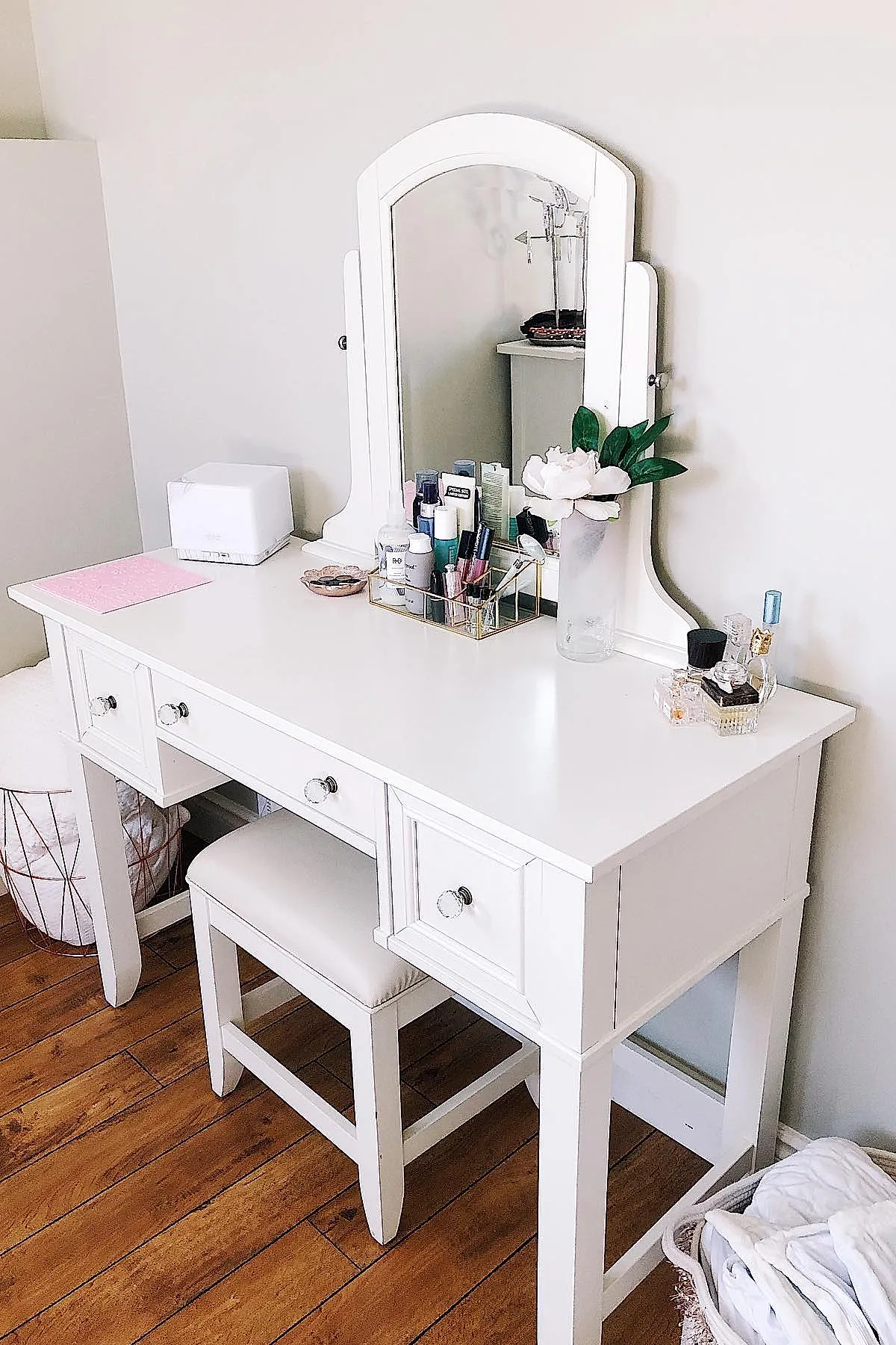 white wooden vanity desk with arched mirror, clear acrylic organizer holding makeup and skincare bottles, white cushioned stool underneath, glass vase with white flower on top, wood floor