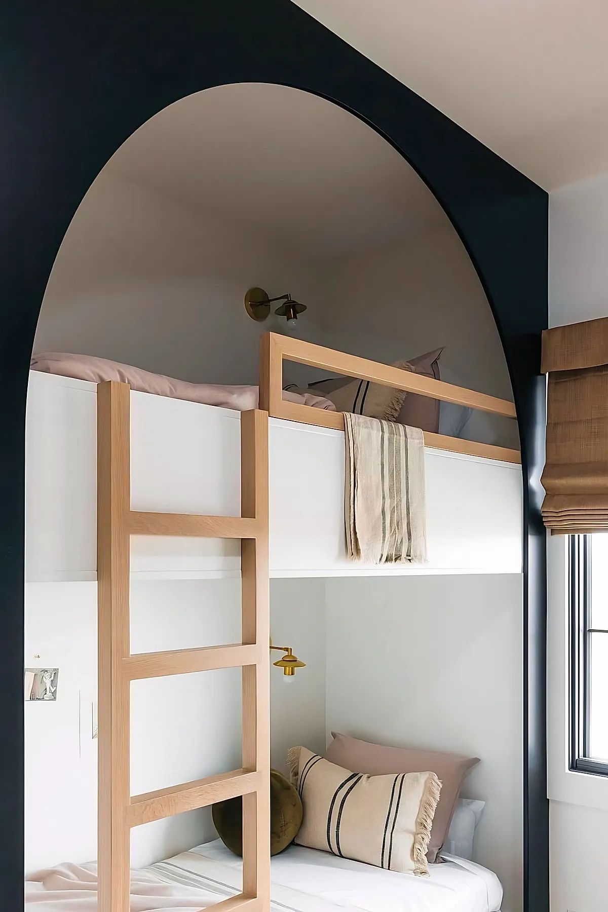 Minimalist adult bunk bed with light wooden frame and ladder, an arched black wall feature around the upper bunk, neutral bedding with striped pillows, brass wall-mounted reading lamp, and a window with woven shades.