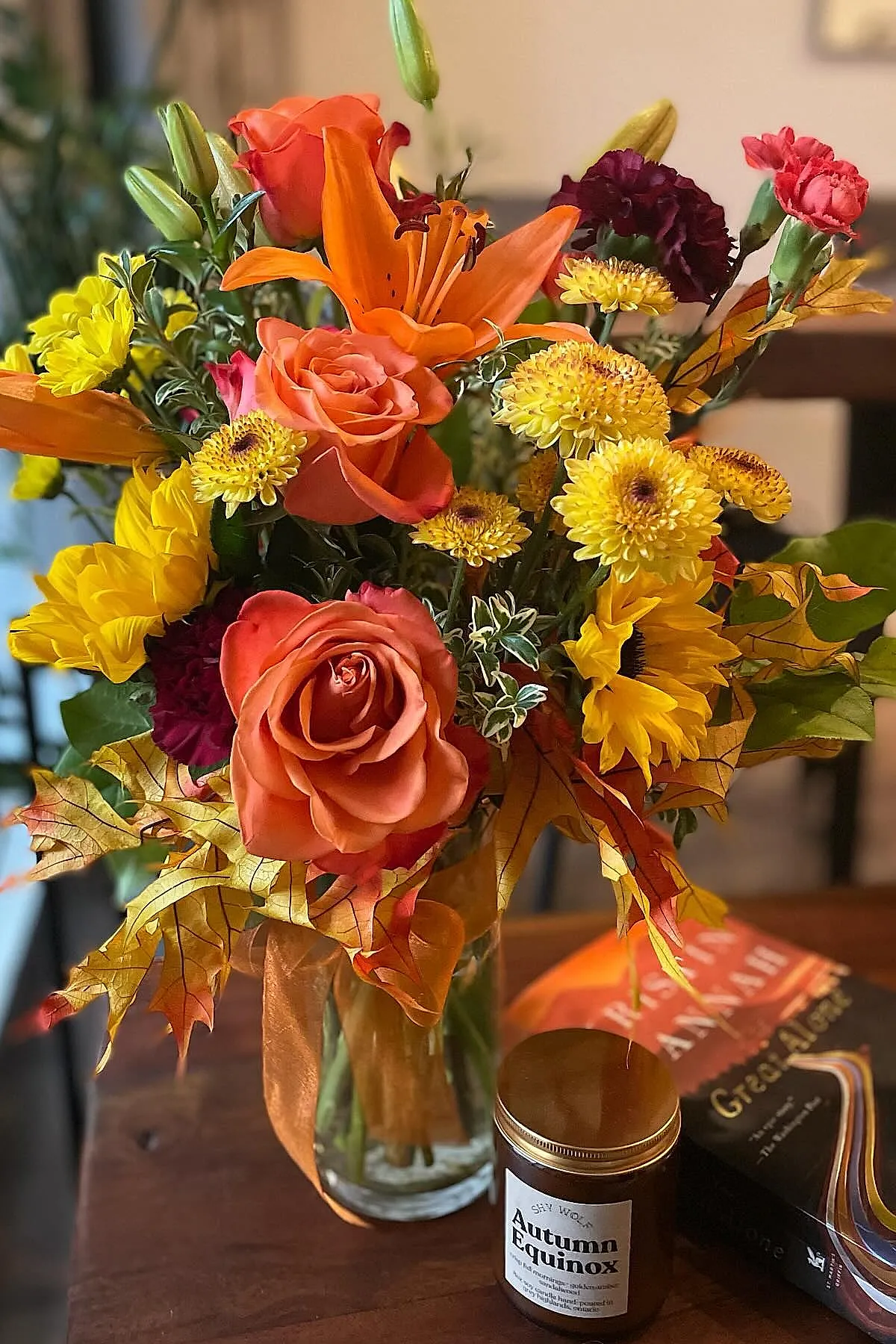 A glass vase filled with bright orange roses, yellow chrysanthemums, sunflowers, burgundy carnations, and orange maple leaves on a wooden table next to a candle labeled Autumn Equinox and books.