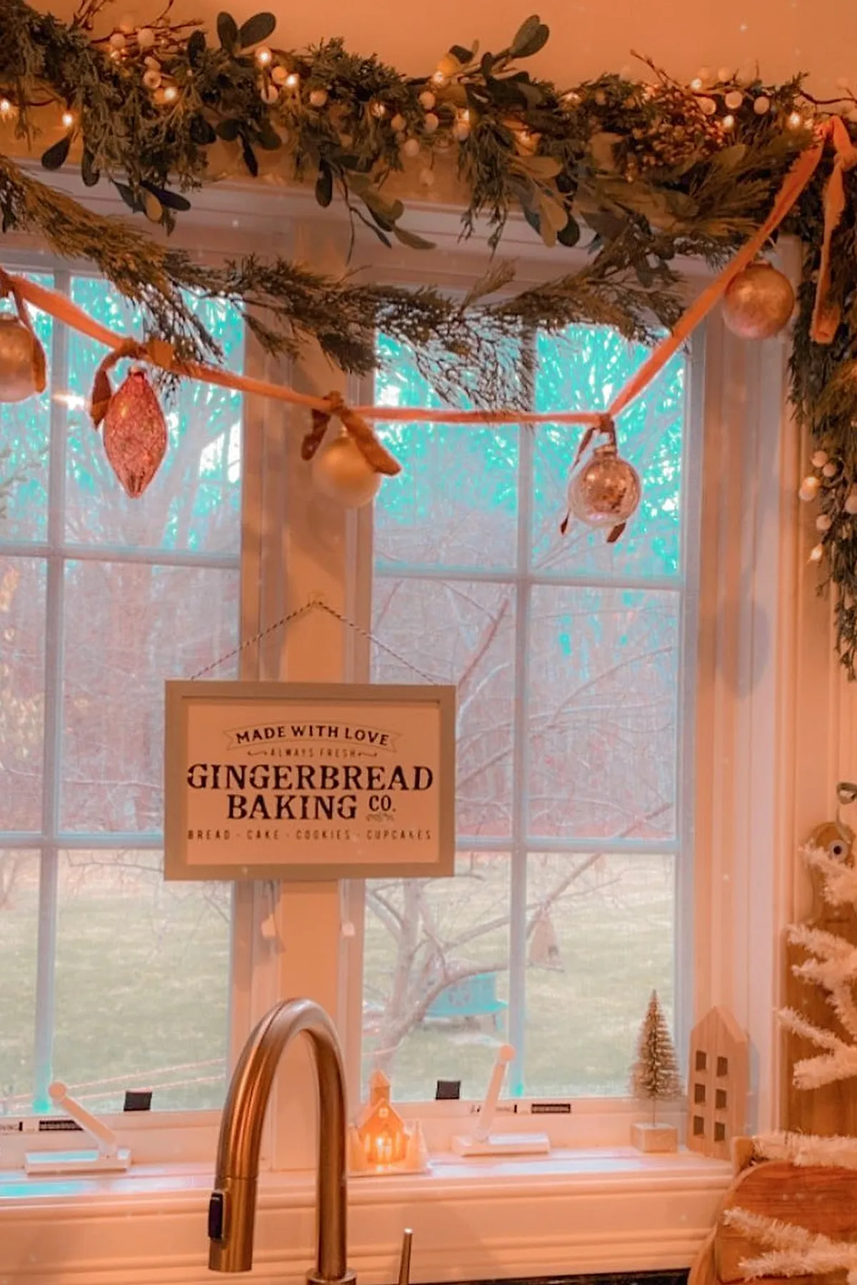 christmas window decorations featuring green pine garlands with white berries and warm string lights above white-framed kitchen windows; hanging ornaments on orange ribbons and a framed