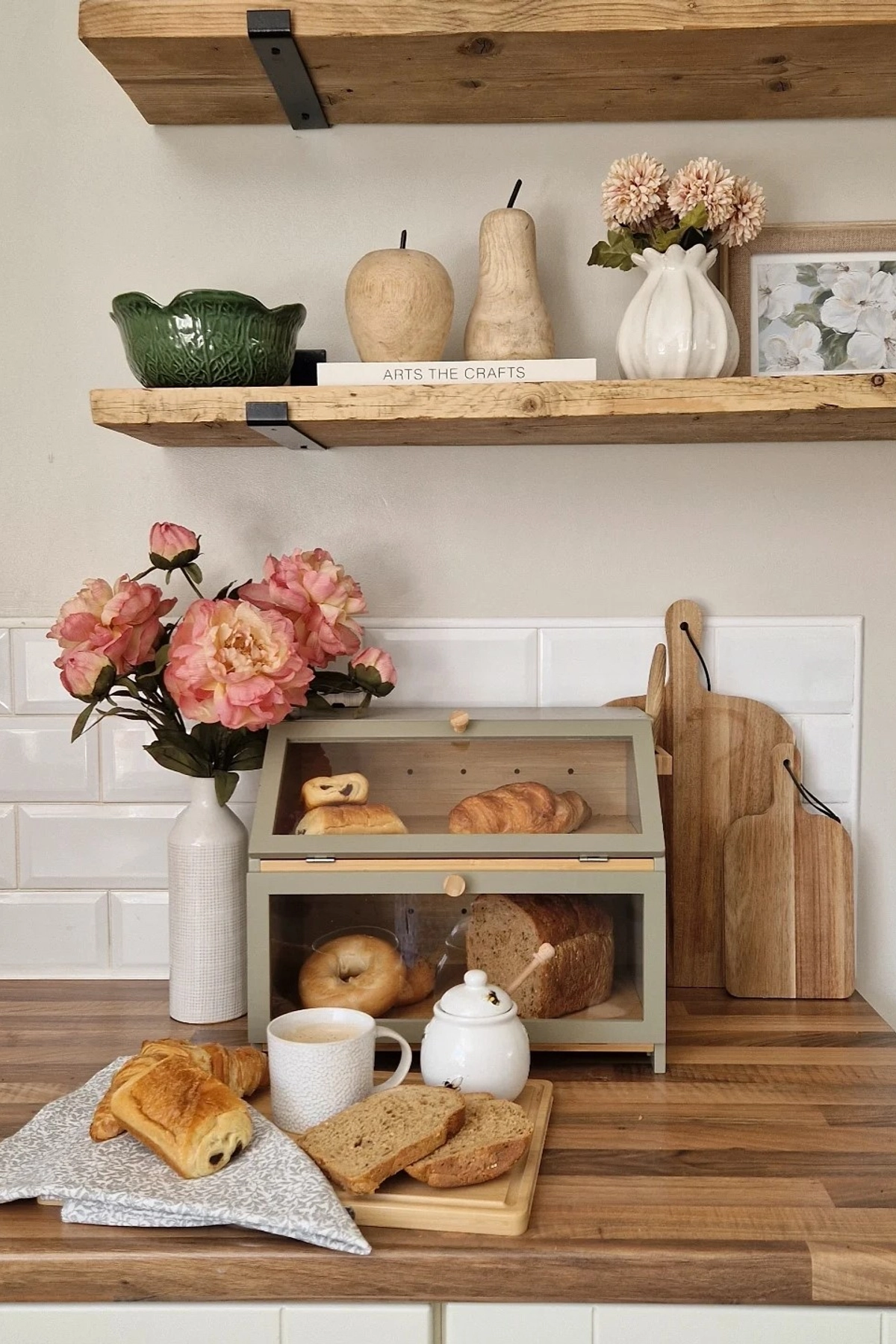 Wooden kitchen countertop with a vase of pink flowers, bread in a box, cutting boards, and pastries on a tray.