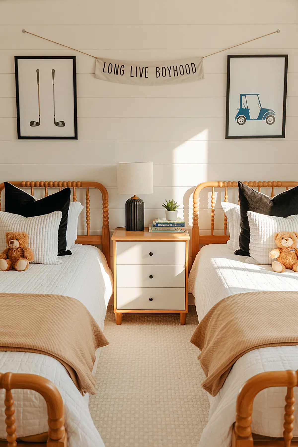 Boys bedroom featuring two wooden spindle twin beds with white quilted bedding, black throw pillows, beige blankets folded at the foot, and small teddy bears on each bed. A nightstand with a dark ribbed lamp and books sits between the beds. White shiplap walls have framed golf clubs and blue golf cart prints flanking a