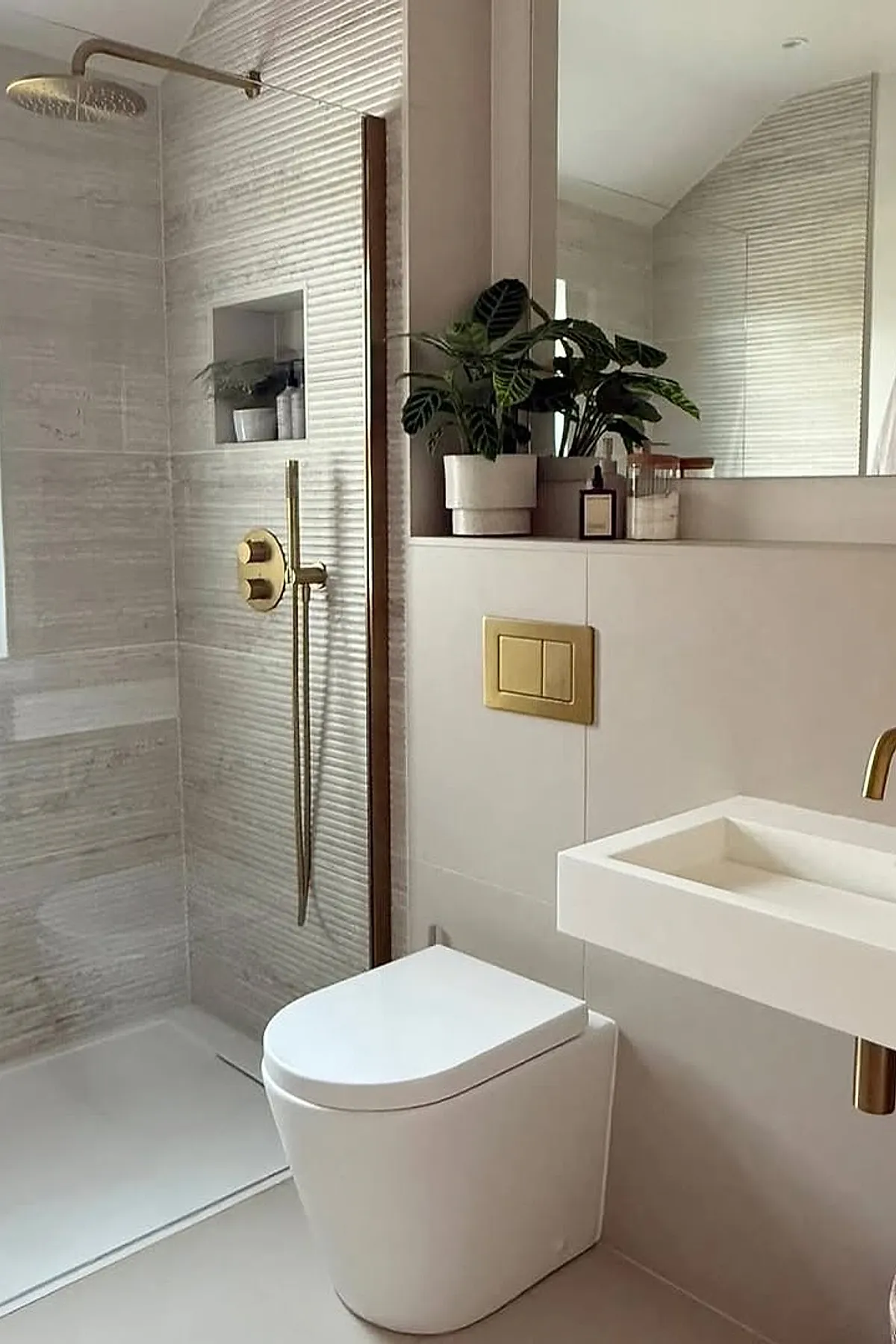 Modern neutral bathroom featuring a walk-in shower with textured beige tiles, brass shower fixtures including a rainfall showerhead and handheld sprayer, a white toilet with concealed cistern, cream-colored wall panels, a floating white rectangular sink with brass faucet, and a large mirror above the sink reflecting the room.