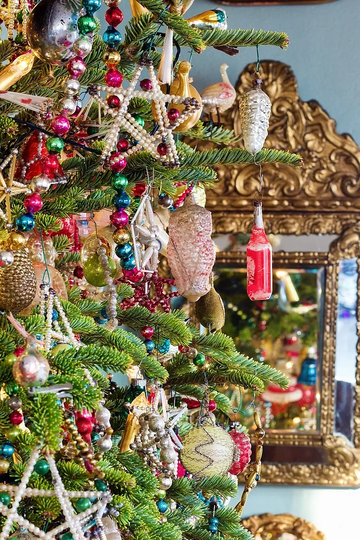 Close-up of a vintage Christmas tree with pearl star ornaments, colorful beaded garlands, and glass decorations shaped like pine cones and bottles, set against an ornate gold-framed mirror on a blue wall.