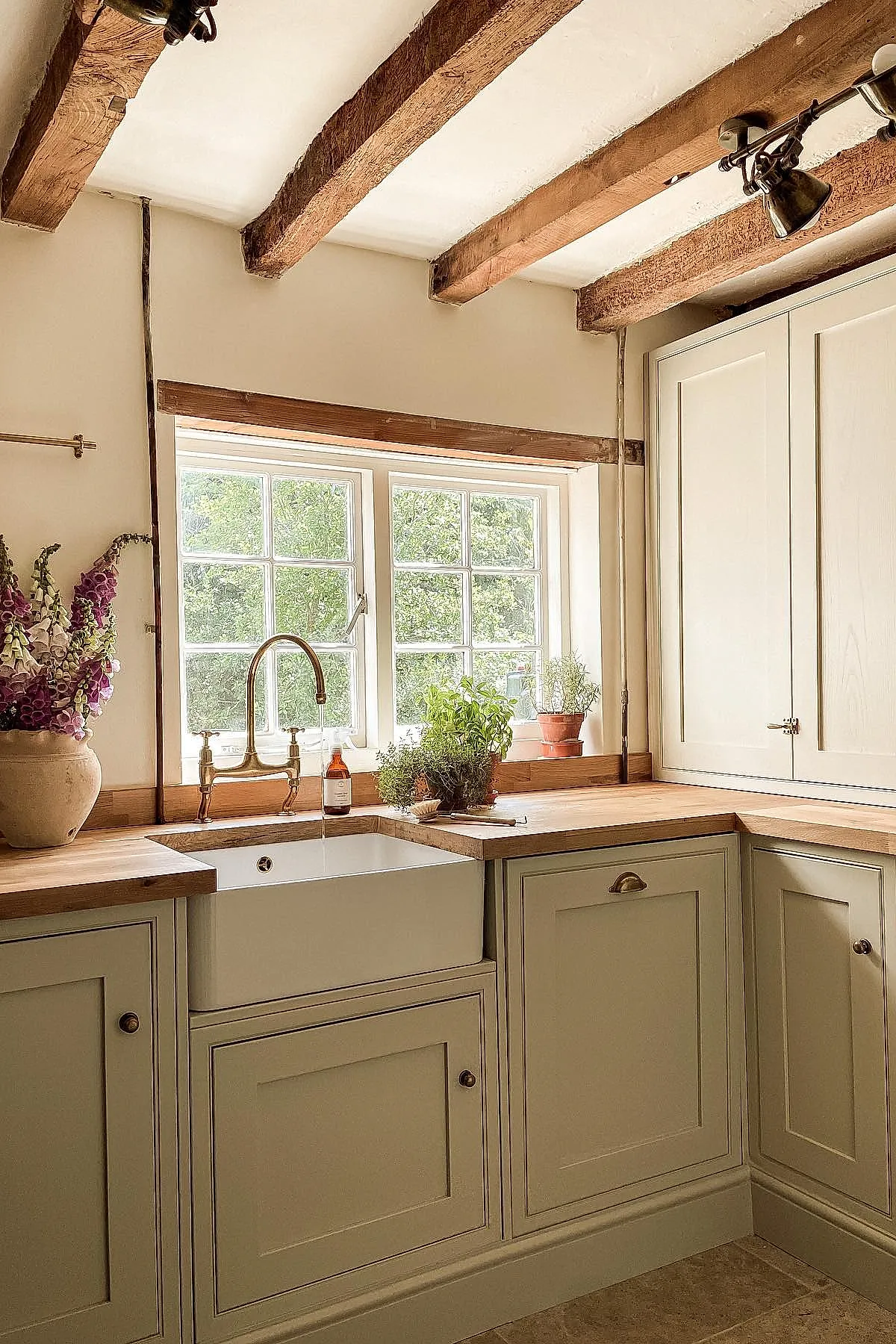 Country cottage kitchen with exposed wooden ceiling beams, cream-colored cabinetry, wooden countertops, farmhouse sink under a window, and potted herbs on the windowsill alongside a vase of purple flowers.
