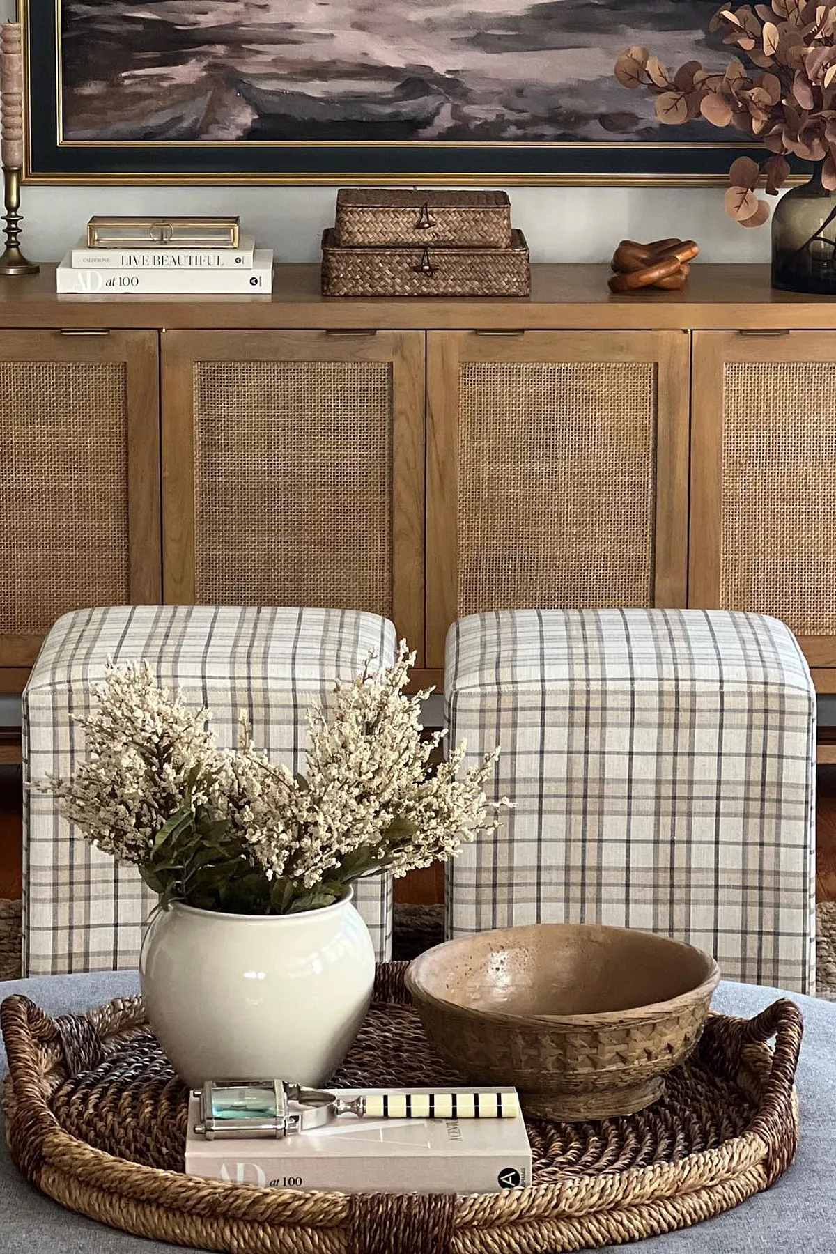 A stylish living space featuring a wooden console table against the wall, decorated with framed landscape artwork, candlesticks, a decorative box, and a vase of dried leaves. In front of the table is a woven tray with a white vase holding fresh flowers, a wooden bowl, and the book TALBOT & CHANDLER. A woven basket sits on the left side of the image, while a gray ottoman with a checkered cushion is located to the right. The overall aesthetic is elegant and serene.
