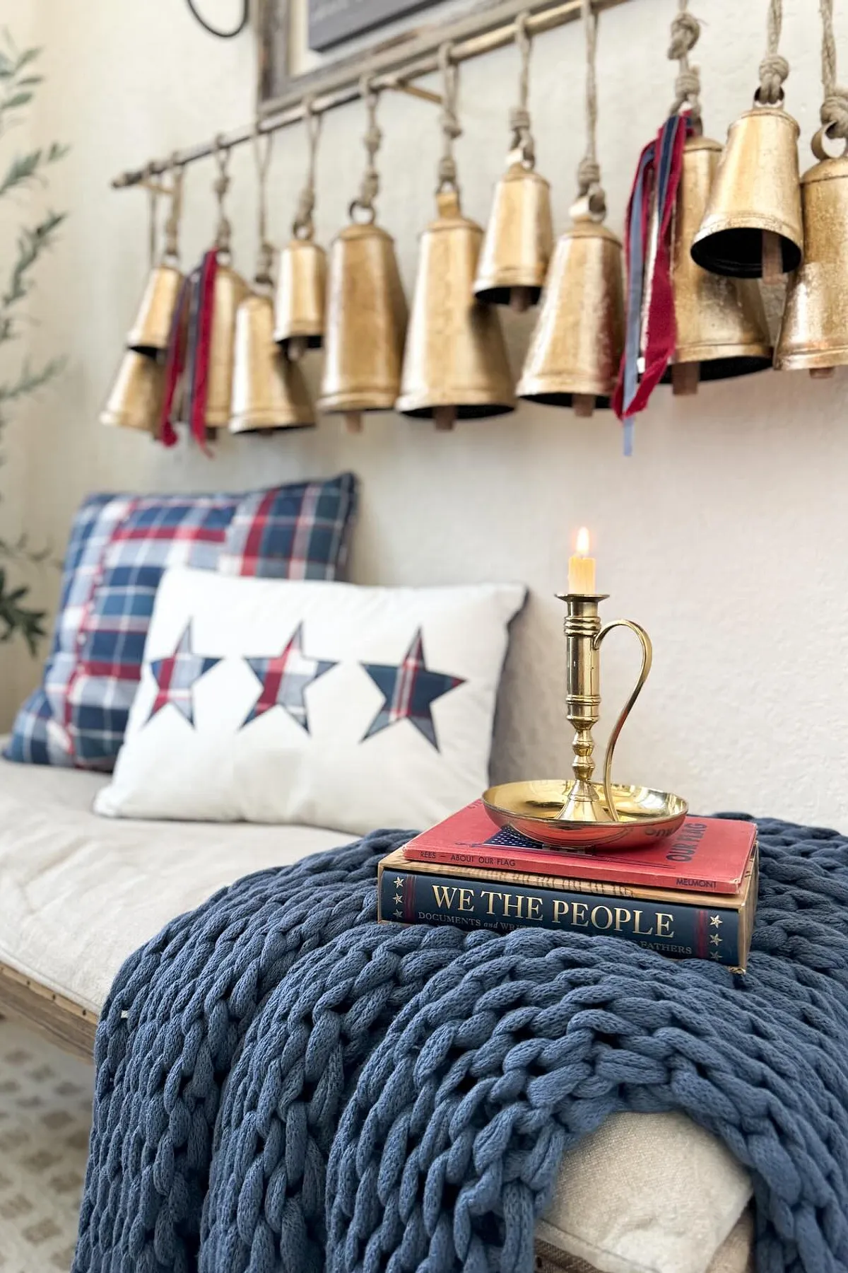 A cozy sofa setup with patriotic pillows, golden bells hanging above, a candle, and a knit blanket.