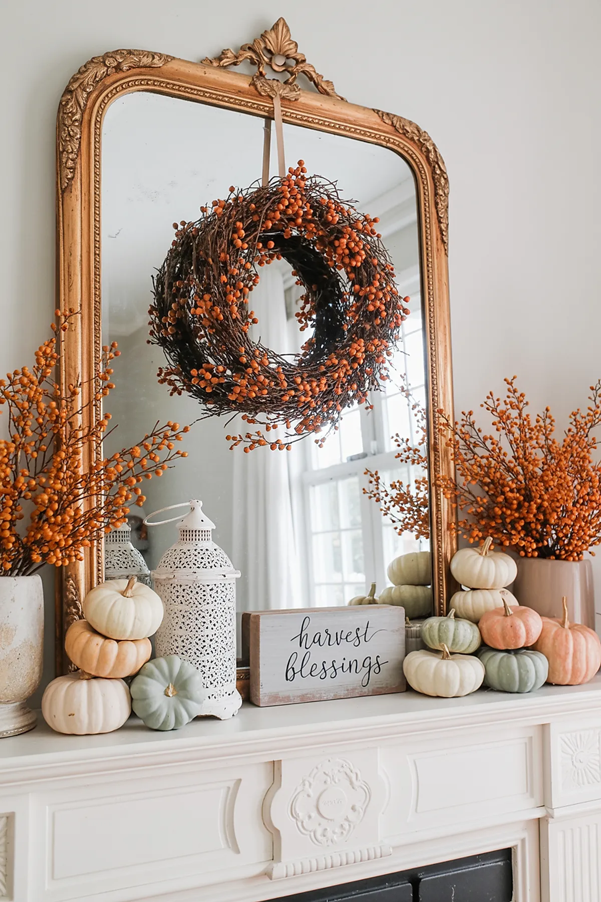 ornate gold-framed mirror above white fireplace mantel decorated with an orange berry wreath hanging from a beige ribbon reflected in the mirror, surrounded by clusters of pale orange, cream, and muted green pumpkins on both sides, two vases filled with matching orange berry branches, and a small wooden harvest blessings sign