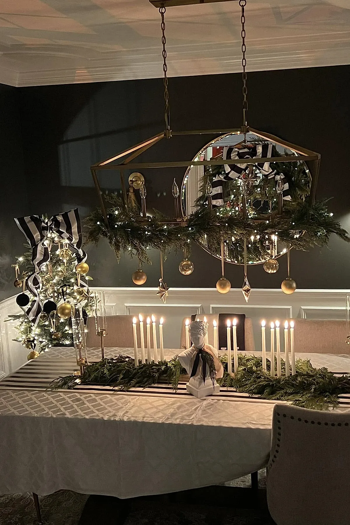 A dining room styled for Ralph Lauren Christmas with a centerpiece of white candles surrounded by greenery, a bust sculpture tied with a black ribbon, a chandelier trimmed with greenery and gold ornaments, and a small tree decorated with striped bows and gold baubles against dark walls.