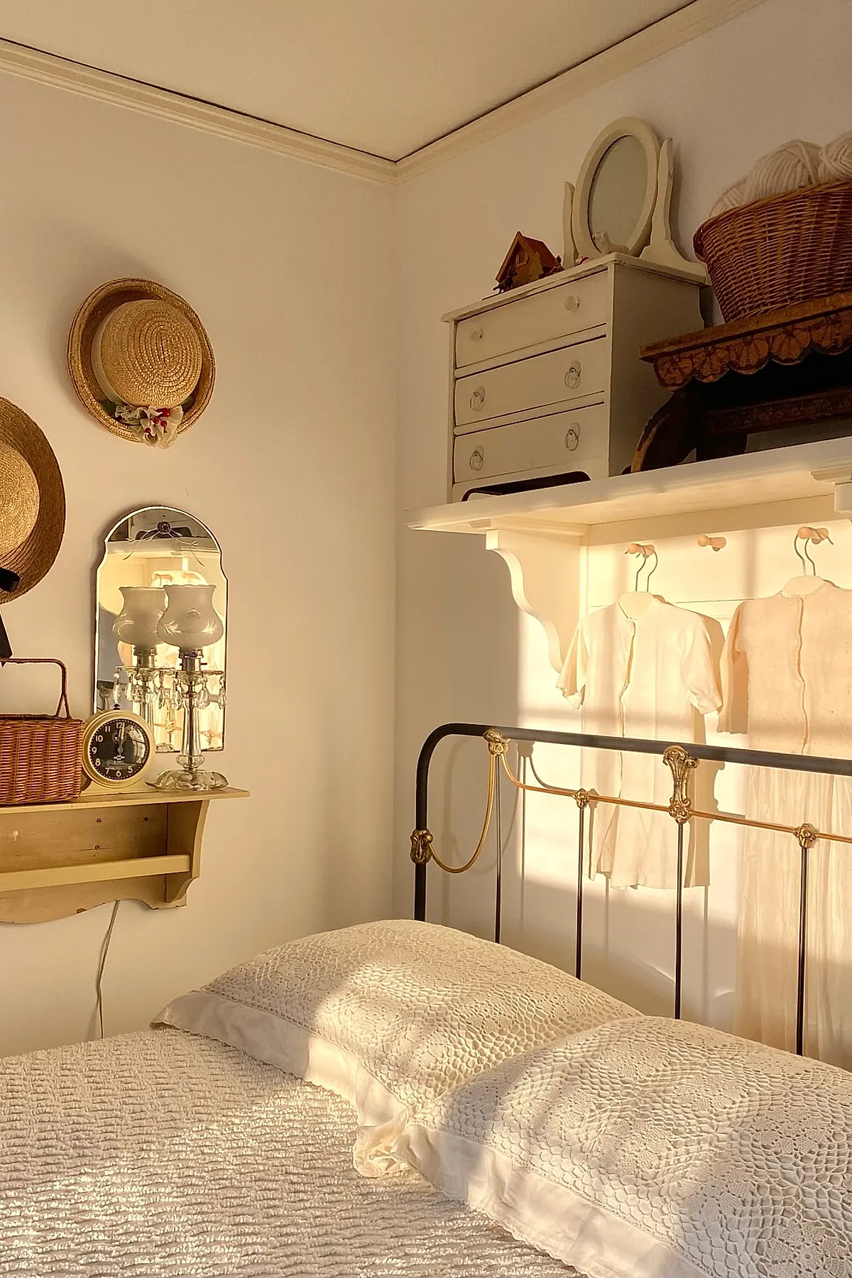 vintage bedroom featuring black iron bed with gold details, white lace quilt and pillows, two straw hats hung on white wall above wooden shelf holding wicker basket and antique clock, small mirror reflecting lamp light