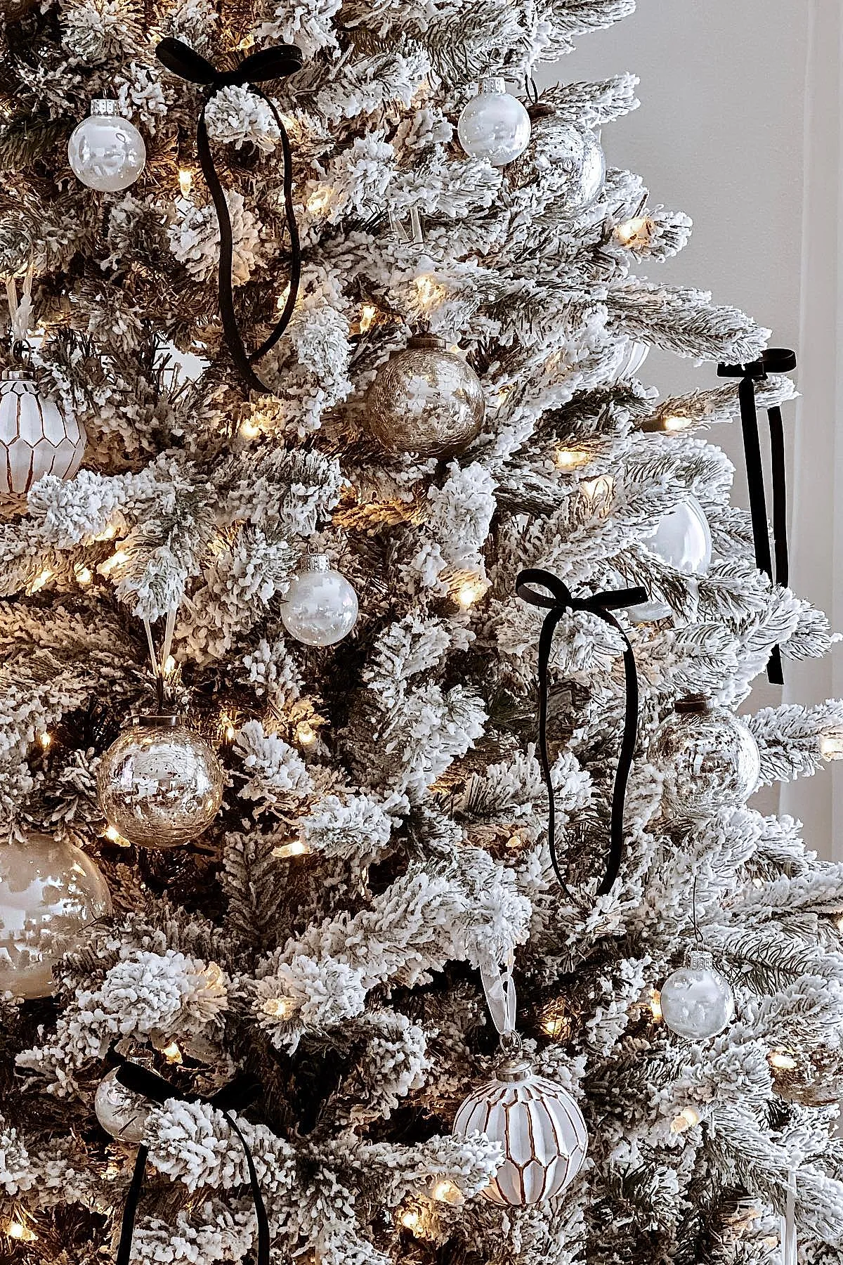 White flocked Christmas tree decorated with clear, white, and silver glass baubles along with black velvet ribbon bows tied on snowy branches lit by warm white lights.