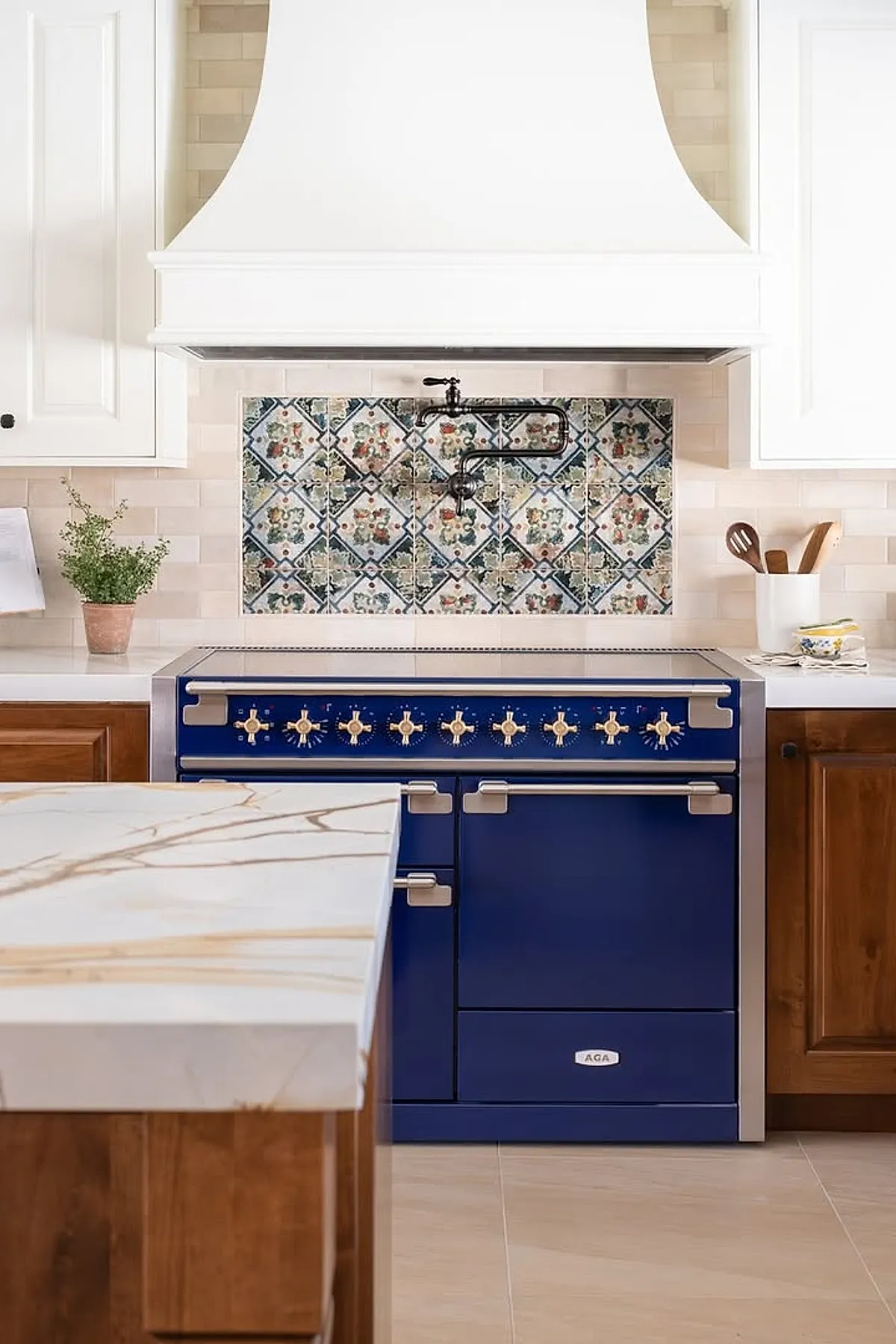 Modern Spanish style kitchen featuring a deep blue AGA stove with brass knobs set against a patterned tile backsplash in blues, greens, and reds, surrounded by white cabinetry and natural wood lower cabinets.