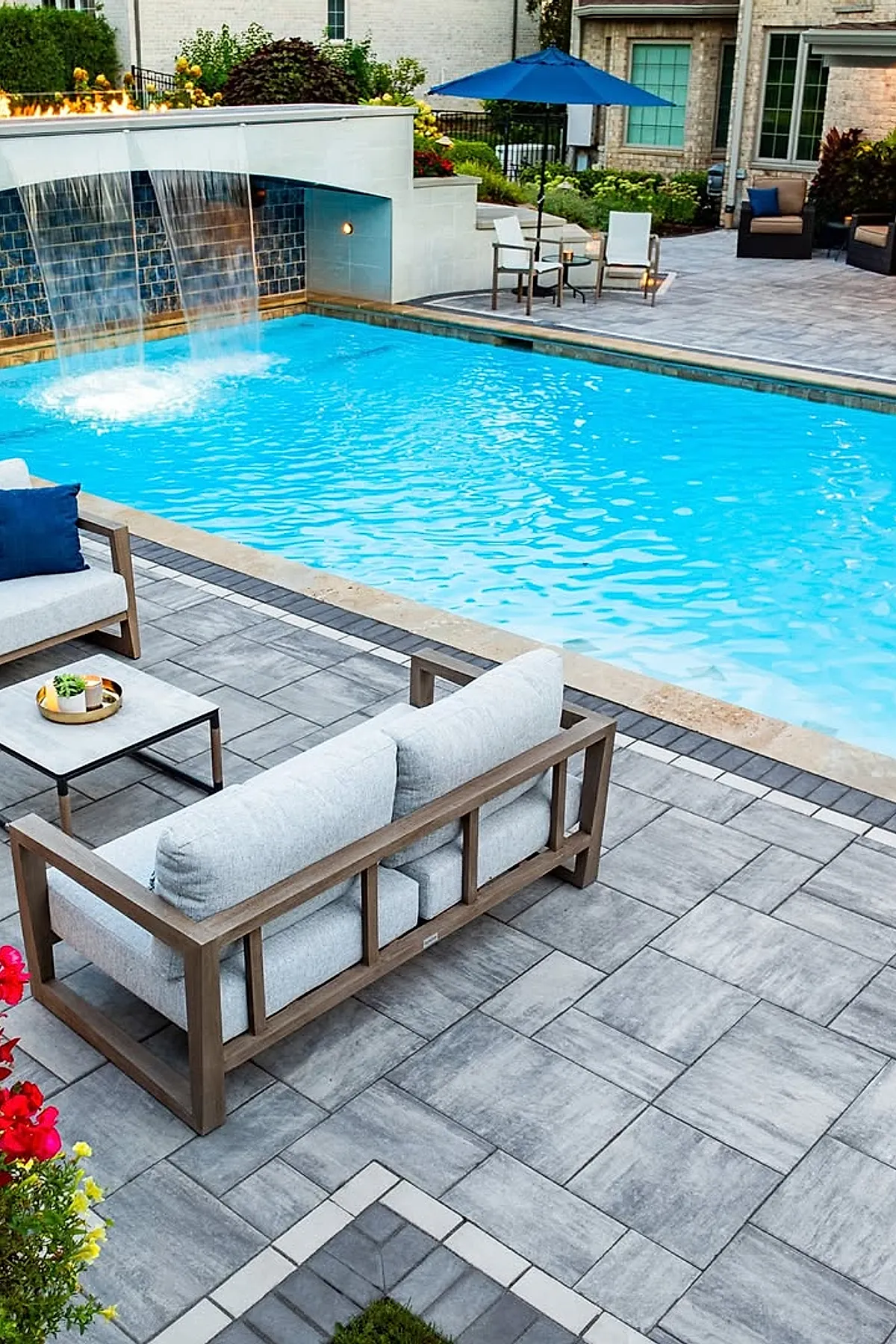 Modern pool deck with gray stone tiles surrounding a bright blue swimming pool featuring a waterfall water feature. The seating area includes light gray cushioned sofas with wooden frames, a small table with plants, and a blue patio umbrella near white chairs, set against a brick house backdrop with greenery.