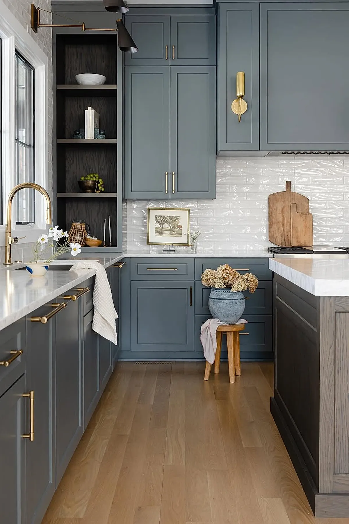 Kitchen with blue-gray cabinetry featuring gold handles, white marble countertops, a wooden stool holding a large blue vase filled with dried flowers, textured white tile backsplash, and wooden cutting boards leaning against the wall.