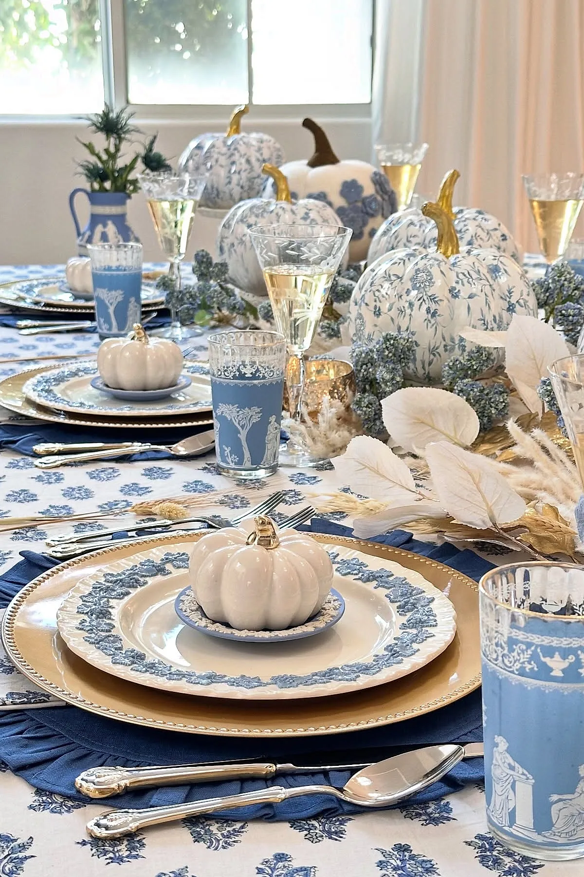 Thanksgiving table with blue and white patterned plates, matching glasses, decorative white pumpkins with blue floral designs and gold stems, set on a floral blue and white tablecloth with silverware and navy blue napkins