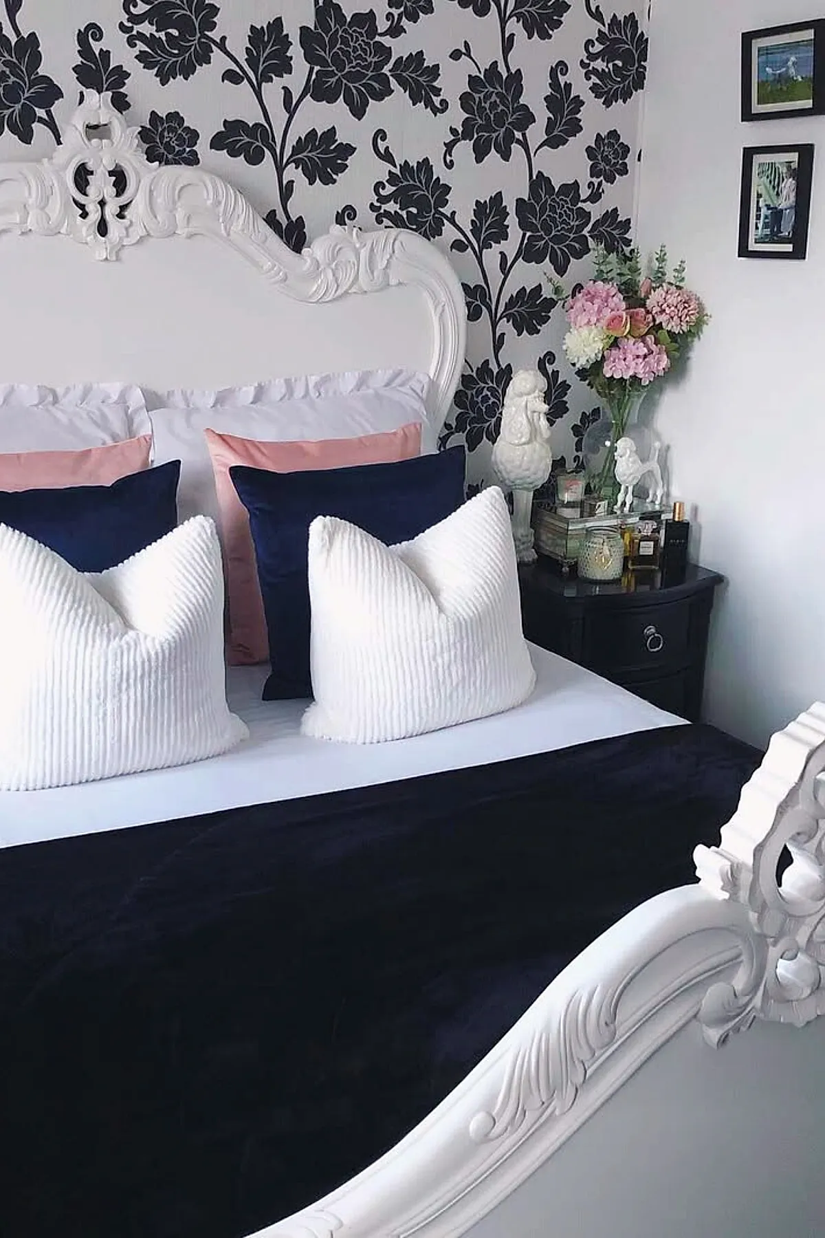 French blue bedroom featuring a white ornate headboard, navy blue pillows, soft pink pillows, large white textured pillows, black floral wallpaper behind the bed, and a black nightstand with decorative objects and fresh pink flowers in a glass vase.