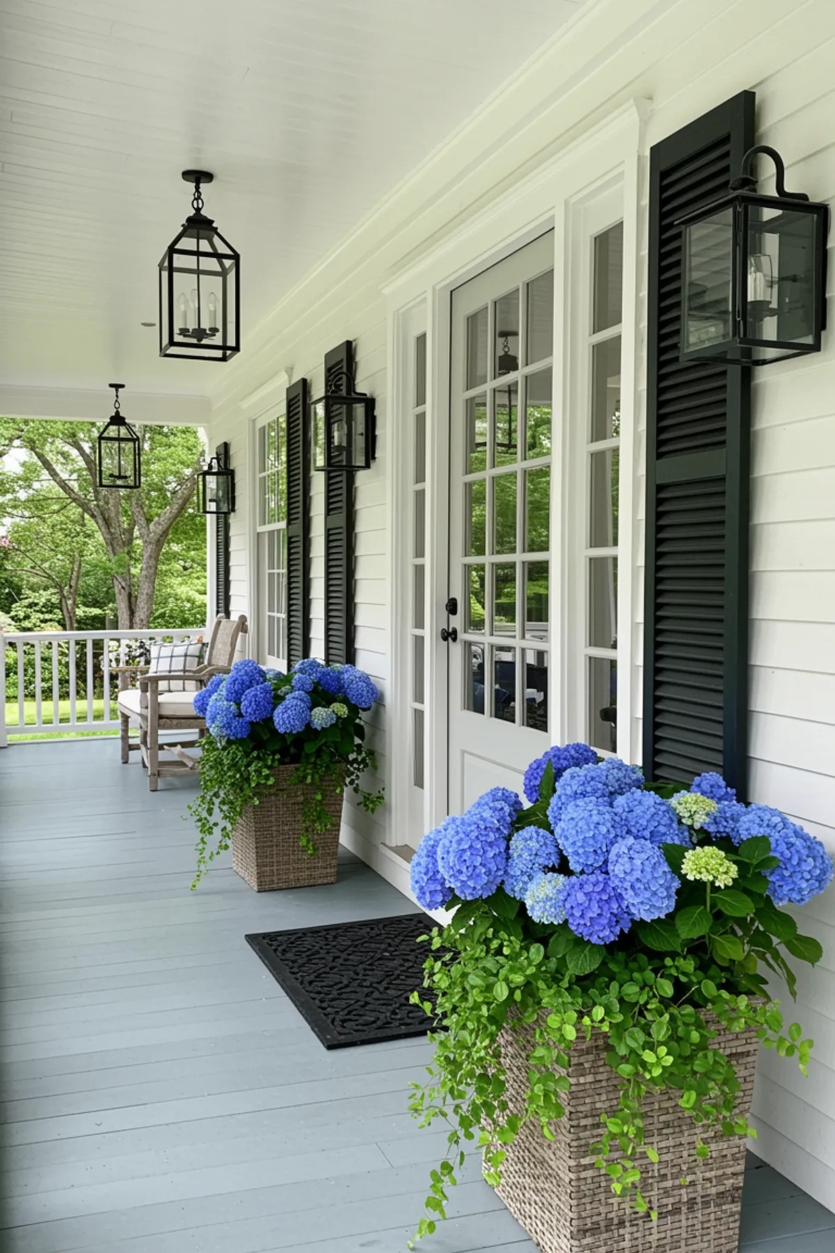 Summer porch showcasing blue hydrangeas in natural planters, black lanterns, and a white door.