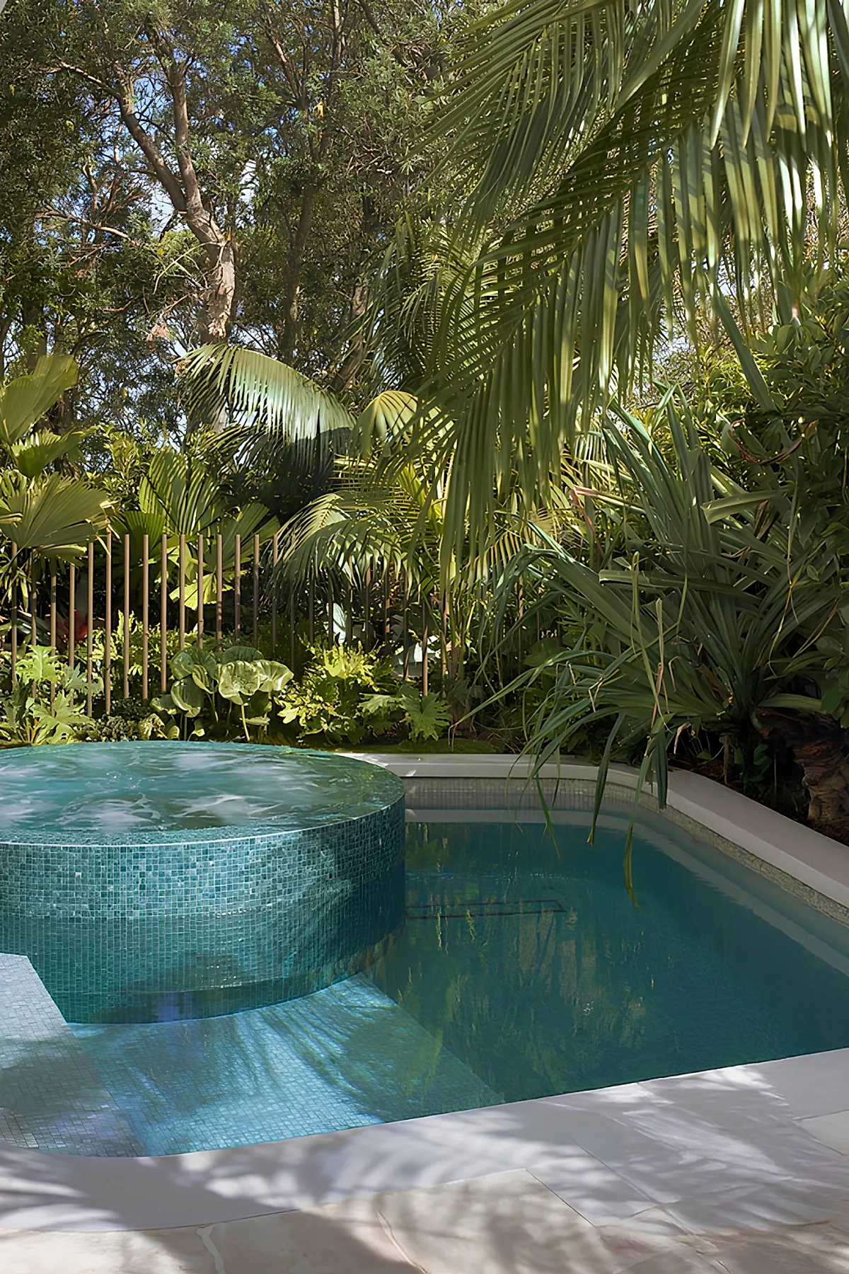Blue-tiled plunge pool with circular elevated spa surrounded by dense tropical greenery including palm fronds and large leafy plants, with a stone patio edge under dappled sunlight.