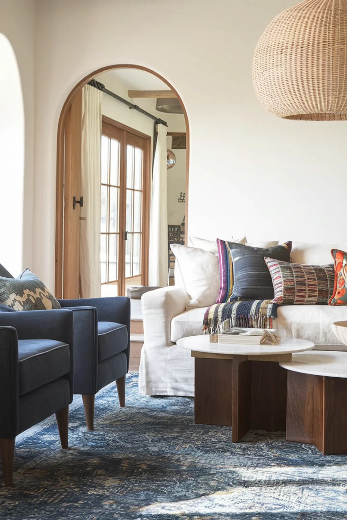Well-lit living room with woven pendant light, blue armchairs, white sofa with colorful pillows, and patterned blue rug.