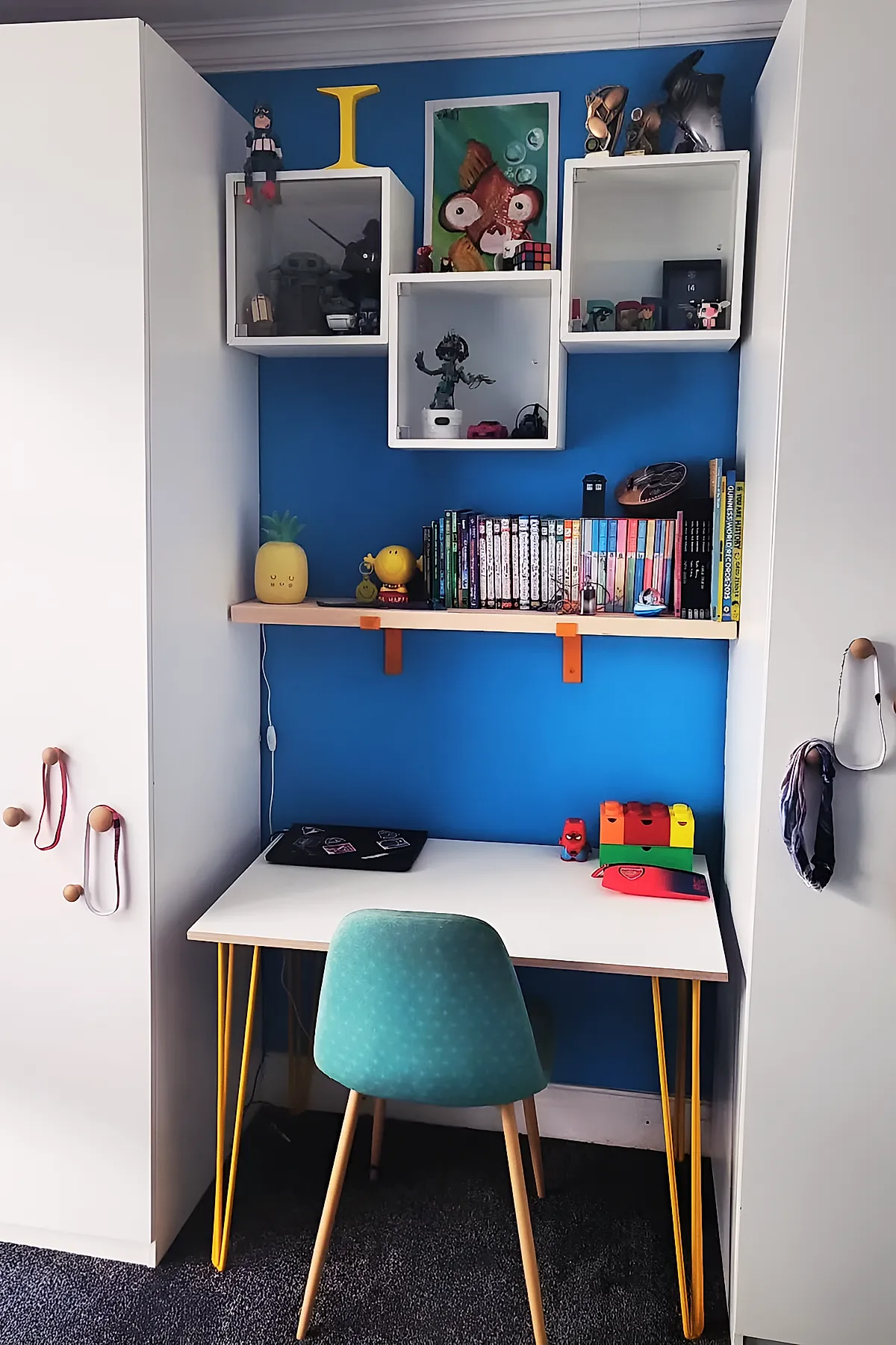 Kids desk area in bedroom featuring a white desk with bright yellow hairpin legs, a teal patterned chair with wooden legs, bright blue accent wall behind the desk, floating wooden shelf holding books and toys, and three white cube shelves mounted above displaying collectibles.