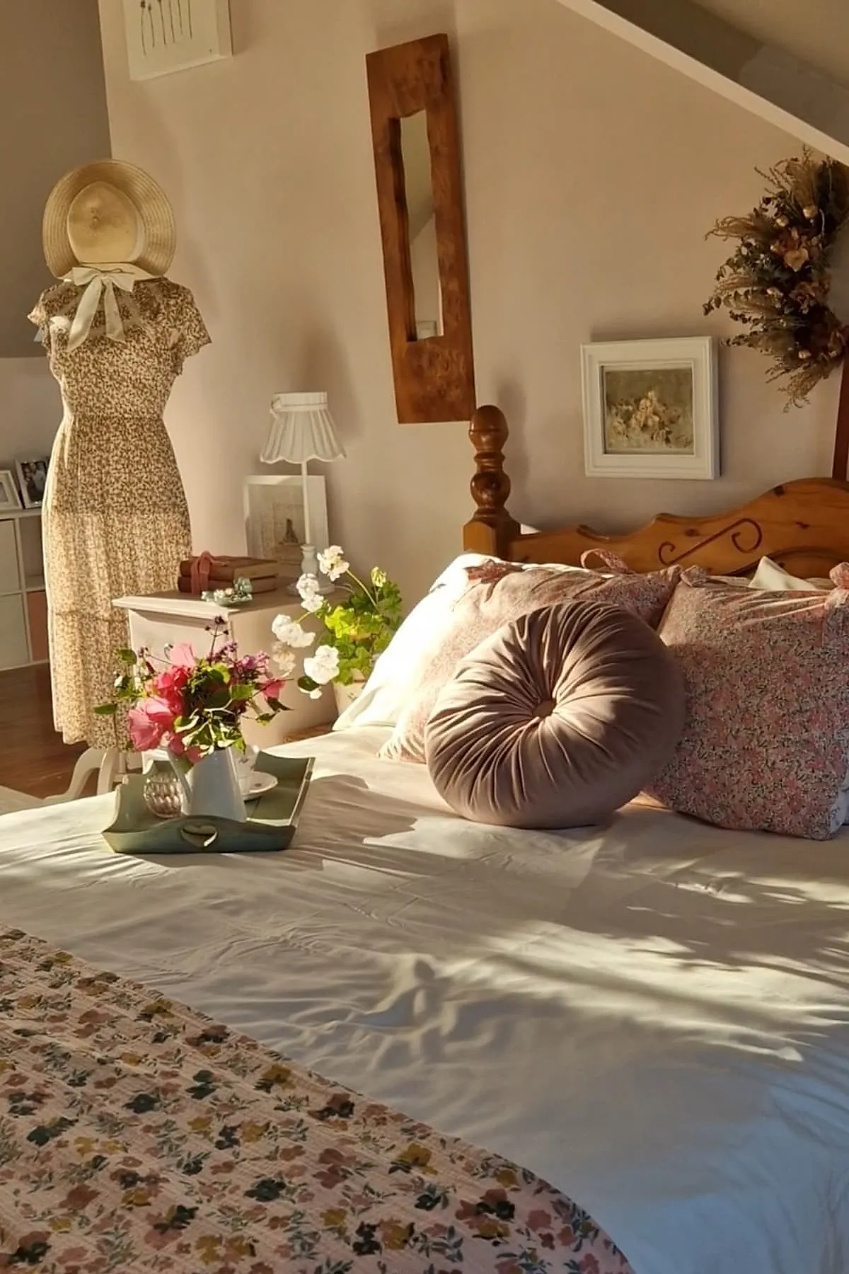 vintage bedroom with wooden bed frame, round blush pink velvet pillow, floral pillows and quilt, vintage dress form wearing beige floral dress with wide-brimmed hat, white nightstand holding lamp and books, dried flower wreath on wall
