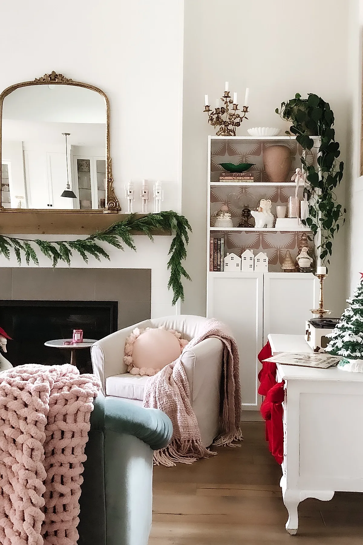 christmas living room featuring a white armchair with pink pom pom pillow and pink knitted throw blanket, green garland draped over wooden mantel above fireplace, large gold framed mirror above mantel, white shelving unit decorated with ornaments and plants, light wood flooring