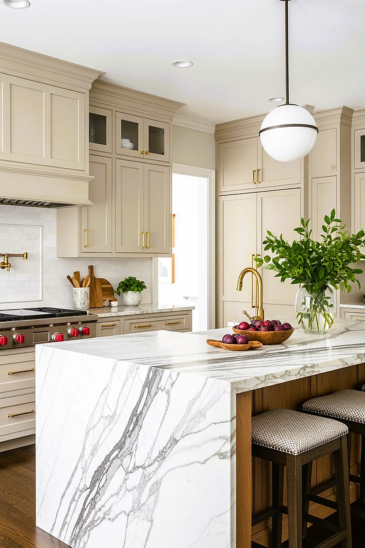 beige kitchen featuring shaker-style beige kitchen cabinets with gold handles, a large white marble waterfall island countertop with gray veining, a gold faucet, dark wooden bar stools with patterned cushions, and a round white pendant light overhead.